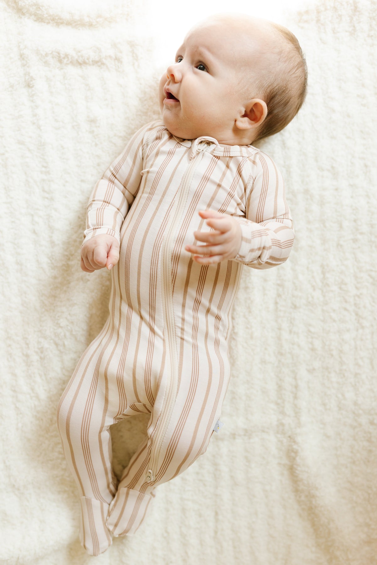 A baby in a toast and honey one piece zipper lies on a soft, white blanket, looking up and smiling.