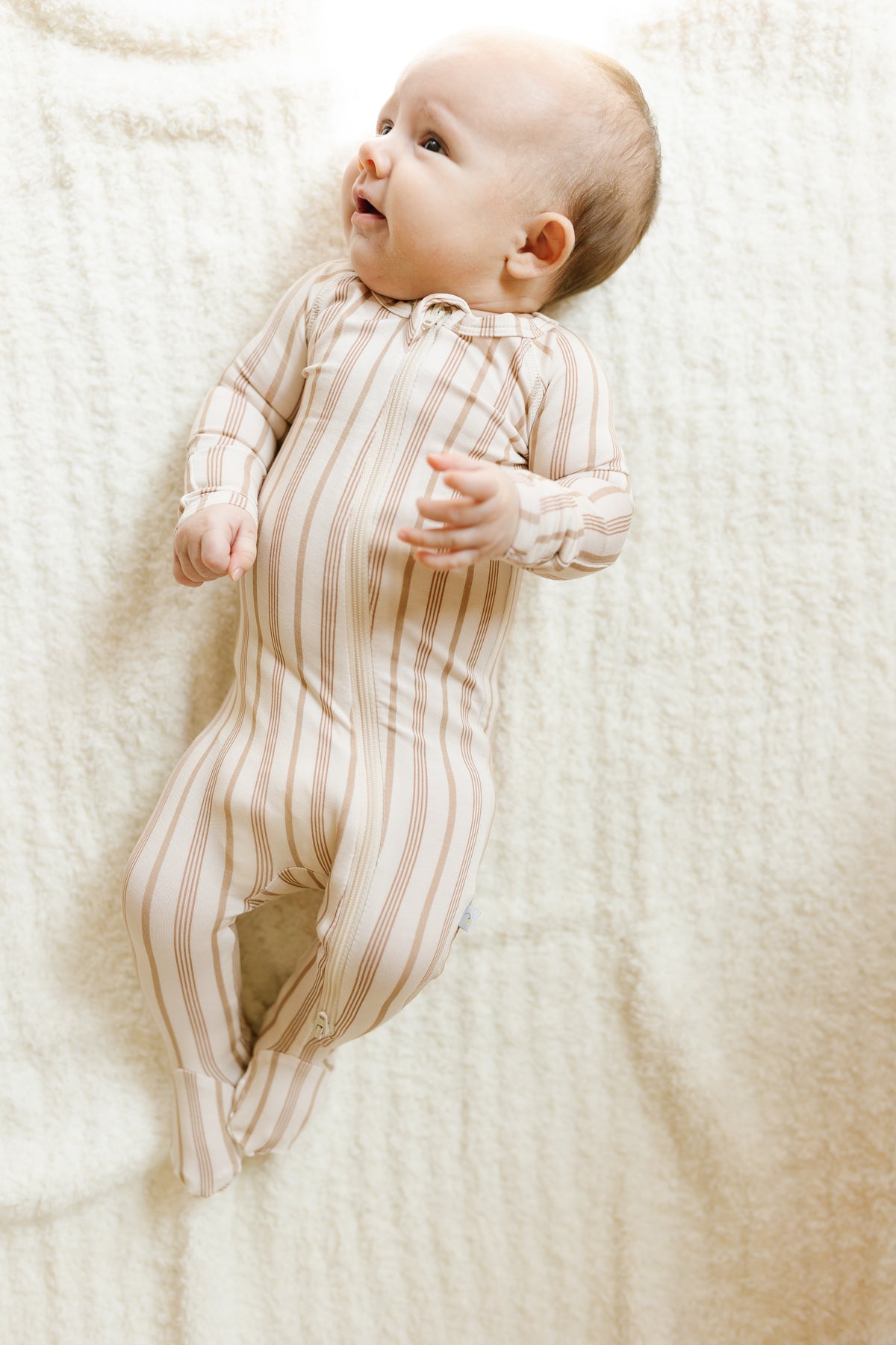 A baby in a toast and honey one piece zipper lies on a soft, white blanket, looking up and smiling.