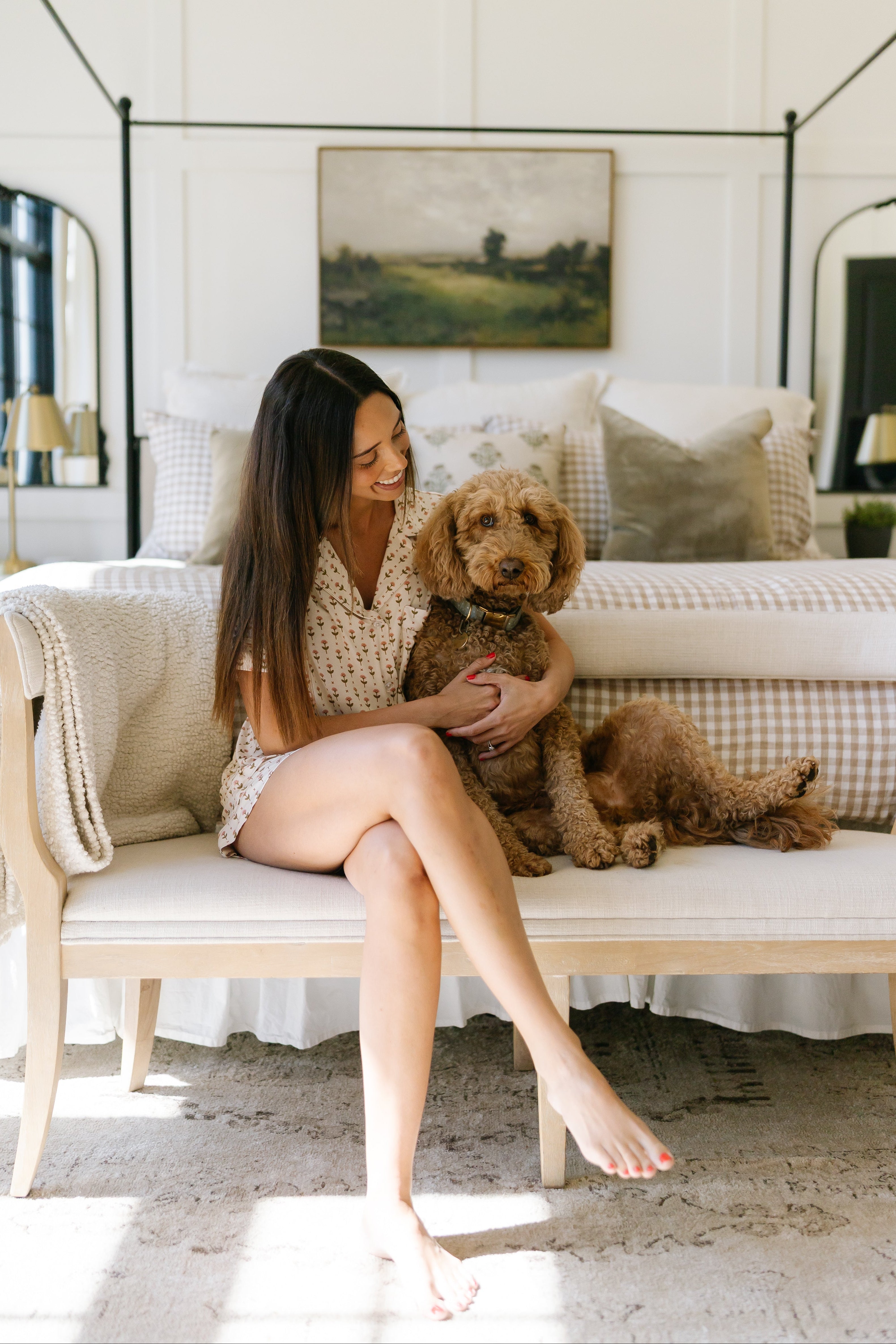 A woman in a tulip pajama set with shorts sits on a bench at the foot of a bed, smiling and hugging a brown curly-haired dog in a bright, cozy bedroom with light decor.