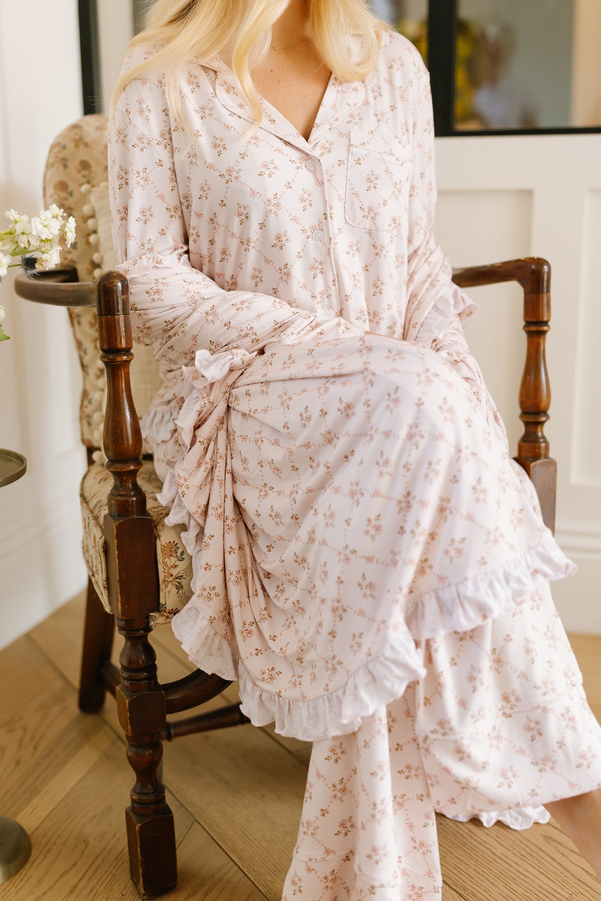 A woman with long blonde hair sits on a vintage chair, wearing the “keepsake / ruffle”—a light-hued, floral-patterned nightgown adorned with delicate ruffles. Her face remains hidden from view. A luxurious blanket is elegantly draped over her lap, and a small table topped with flowers stands nearby.