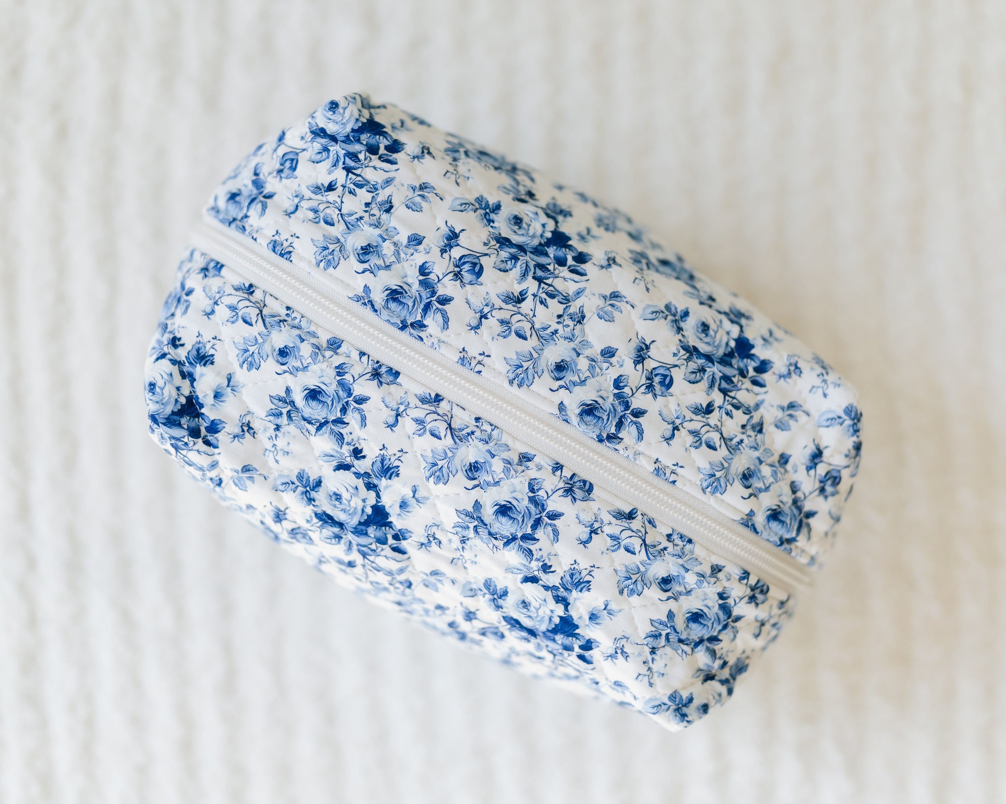 A rectangular fabric pouch with a blue floral pattern and white zipper, the blue haven / travel bag is a travel essential displayed on a textured white surface.