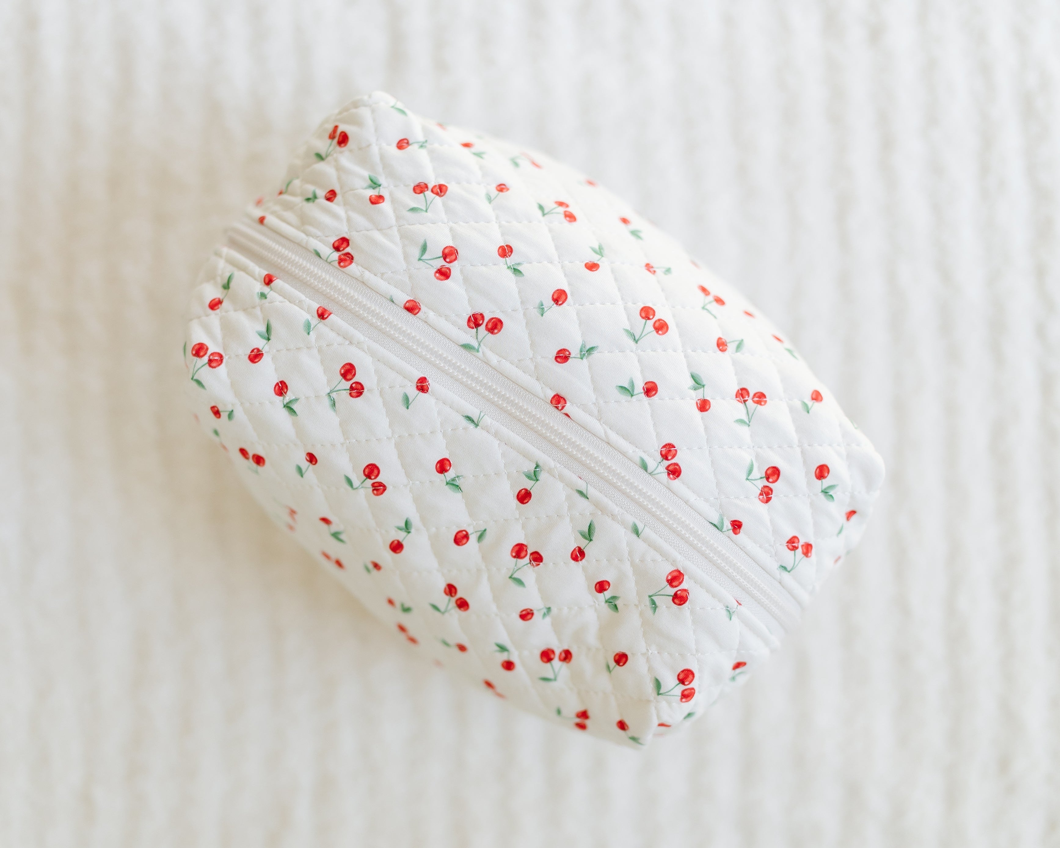 The cherry on top / travel bag is a small, quilted white pouch with a zipper and cute cherry print, shown on a textured white surface—perfect for carrying your essentials.
