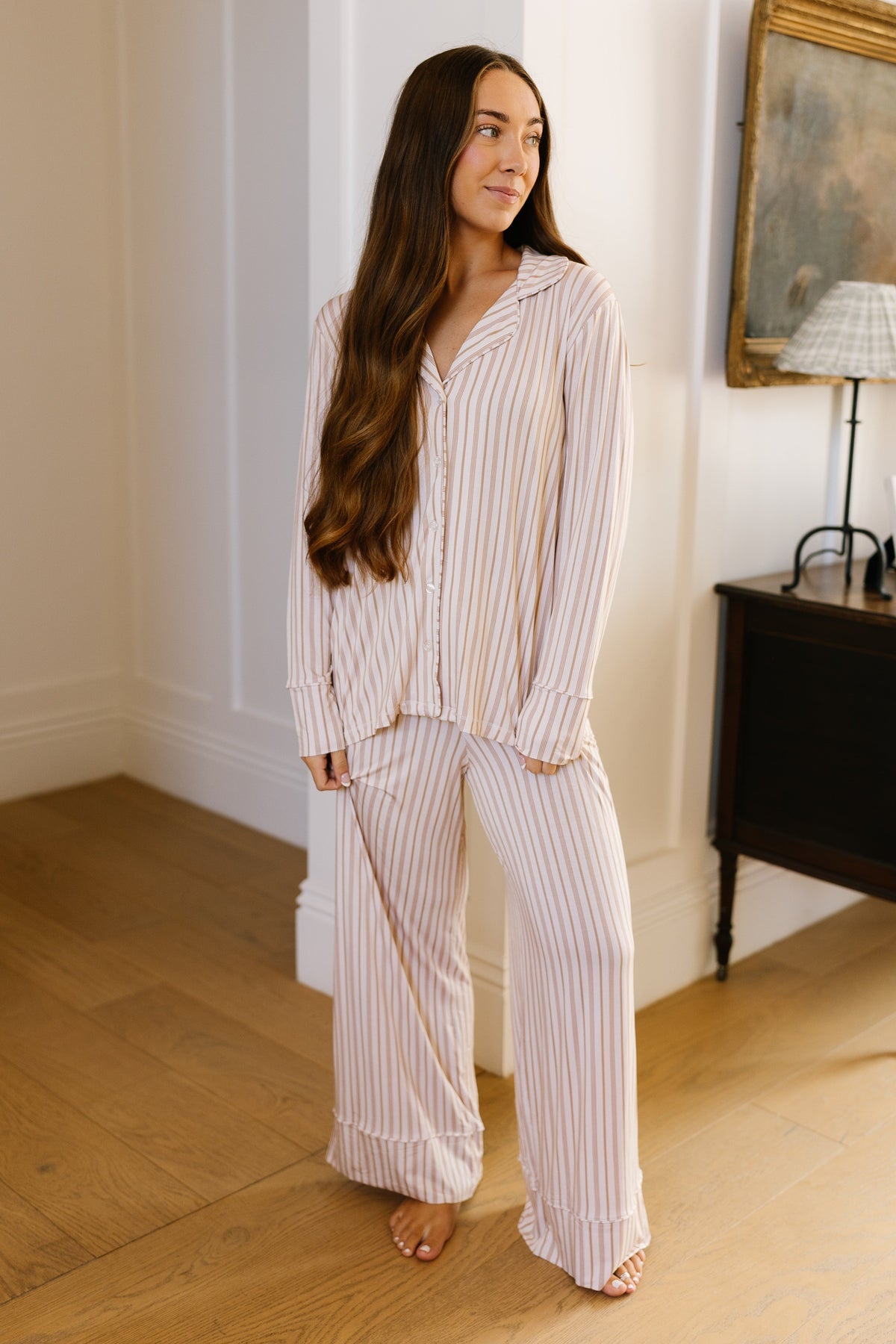 A woman with long brown hair stands indoors, barefoot on a wooden floor beside a dark side table and a lamp, wearing the toast and honey pajama set with long pants—a relaxed fit, light-colored striped loungewear piece that perfectly embodies at-home comfort.