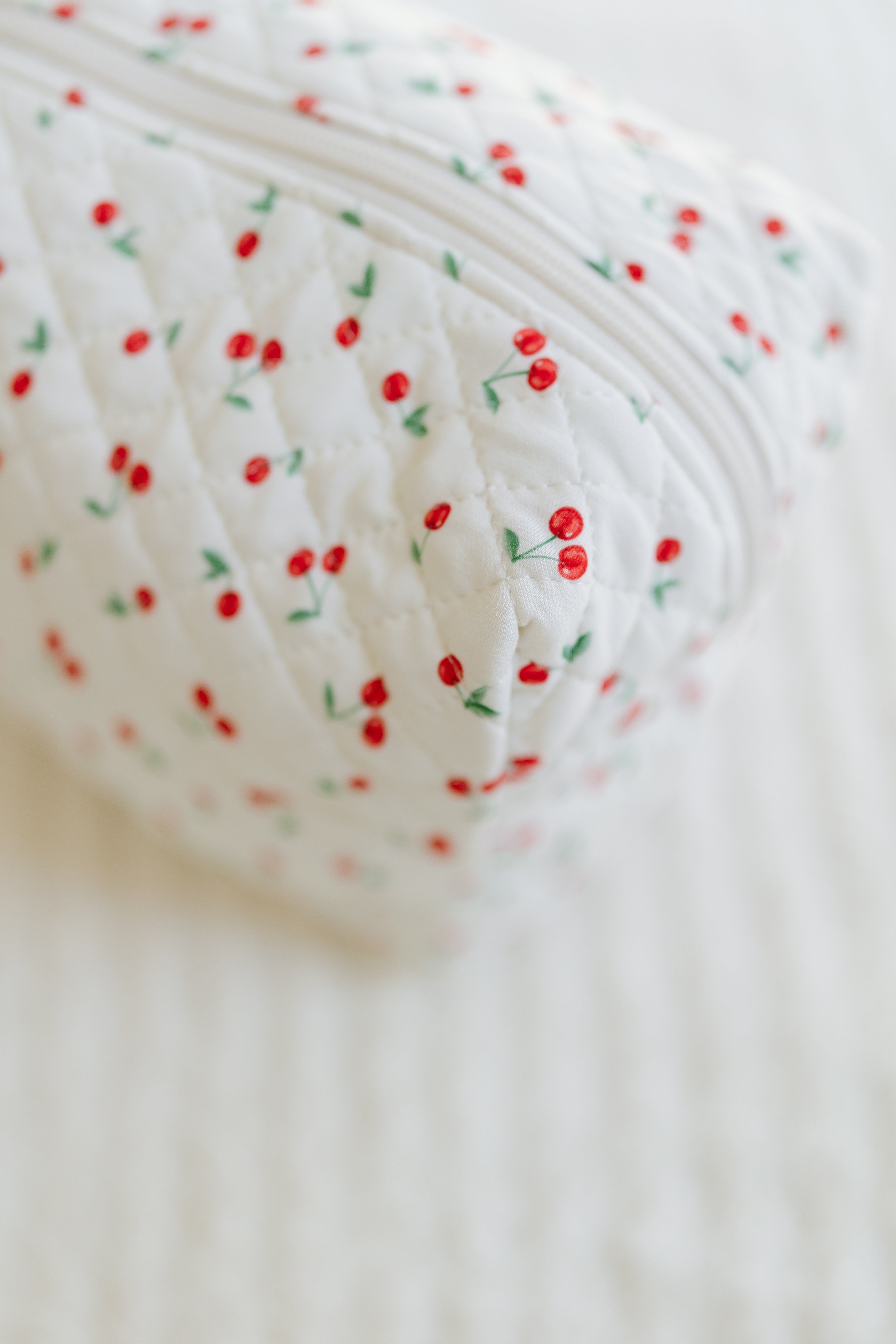 A close-up of the cherry on top / travel bag, made from quilted white bamboo viscose with a zipper and a red cherry print with green stems. The softly textured white background highlights its stylish design.