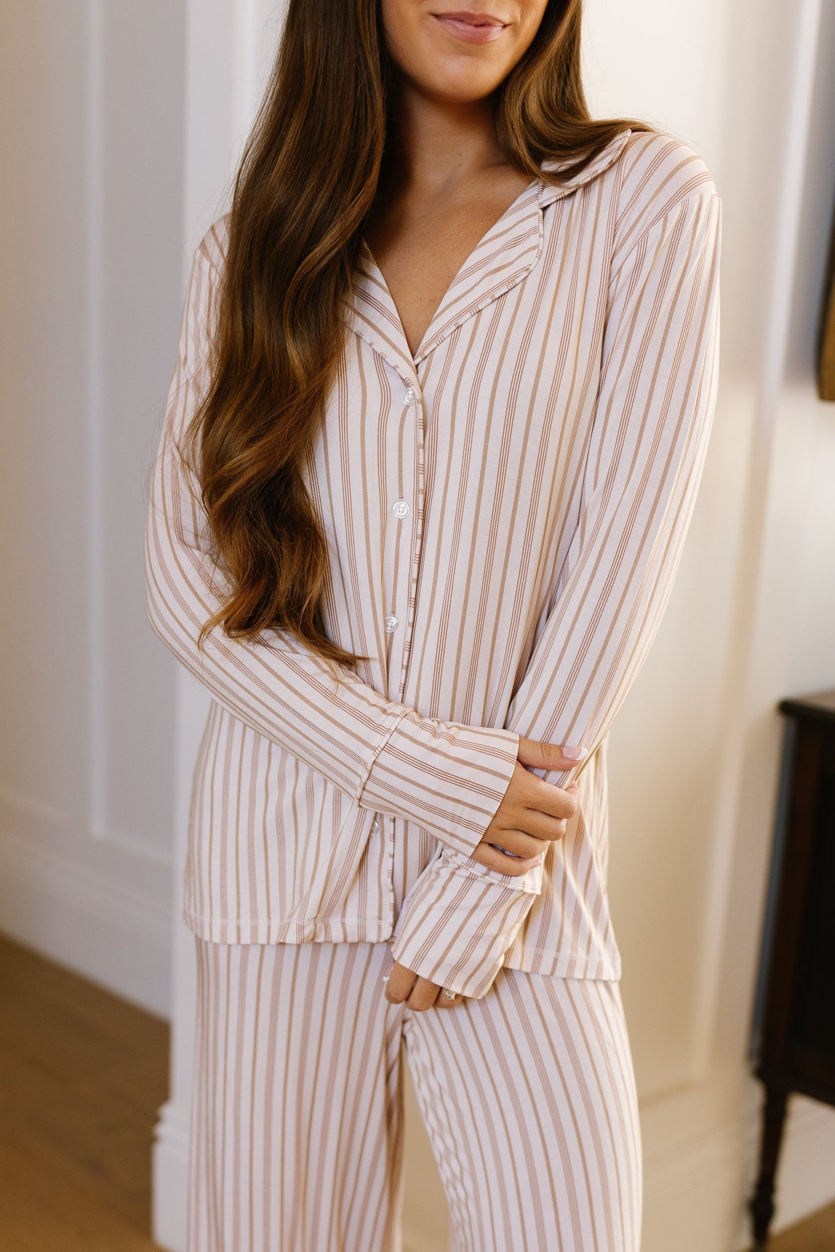 A woman with long brown hair wears the toast and honey / pajama set / long pant in beige and white stripes, standing indoors with her arms relaxed in front of her. Her face is partially visible against a backdrop of a white wall and wooden floor—perfect lounge wear for those who appreciate a relaxed fit.