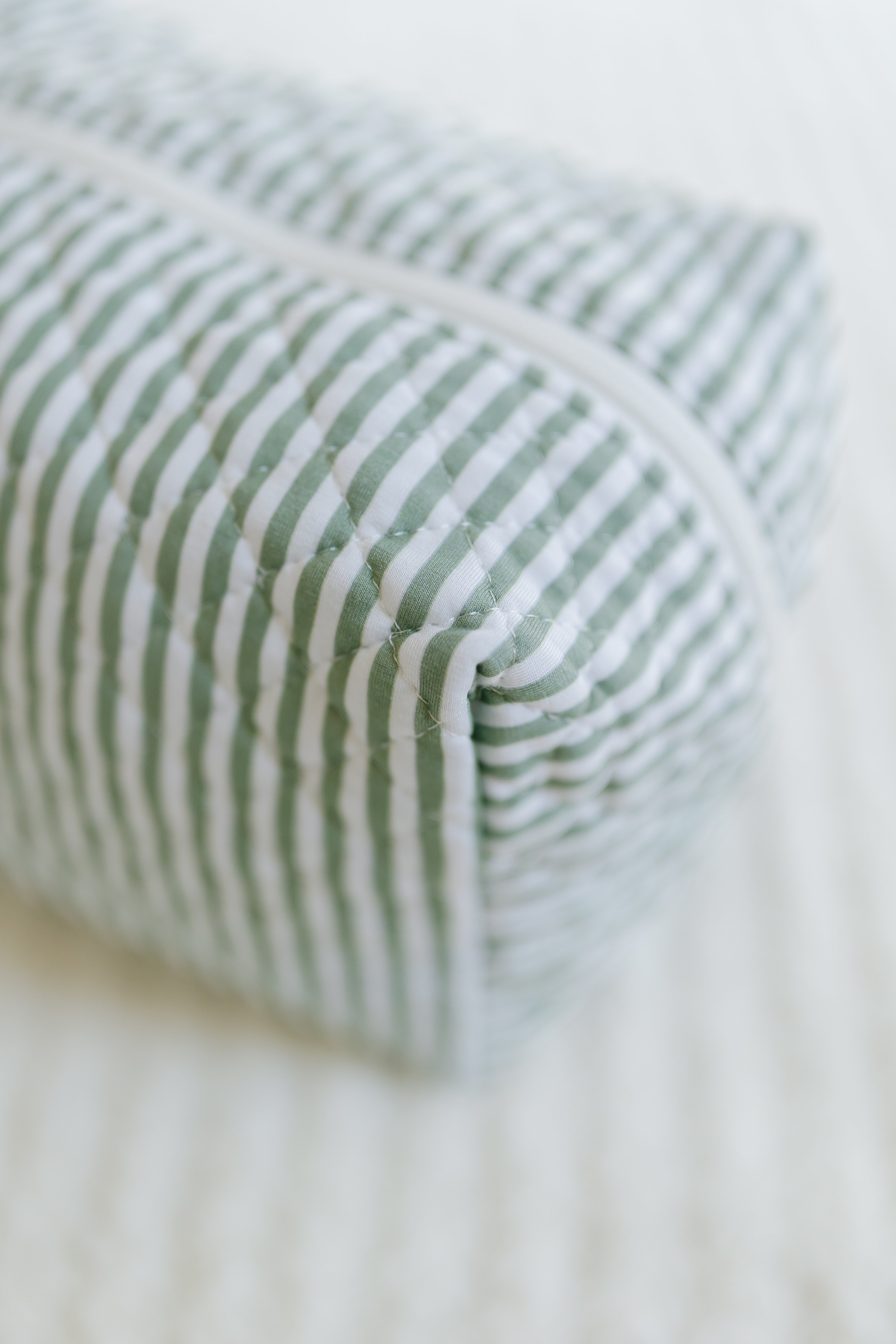 A close-up of the Tree House Travel Bag, a rectangular quilted bag with green and white vertical stripes and a white zipper, resting on a light textured surface—a truly stylish travel accessory.