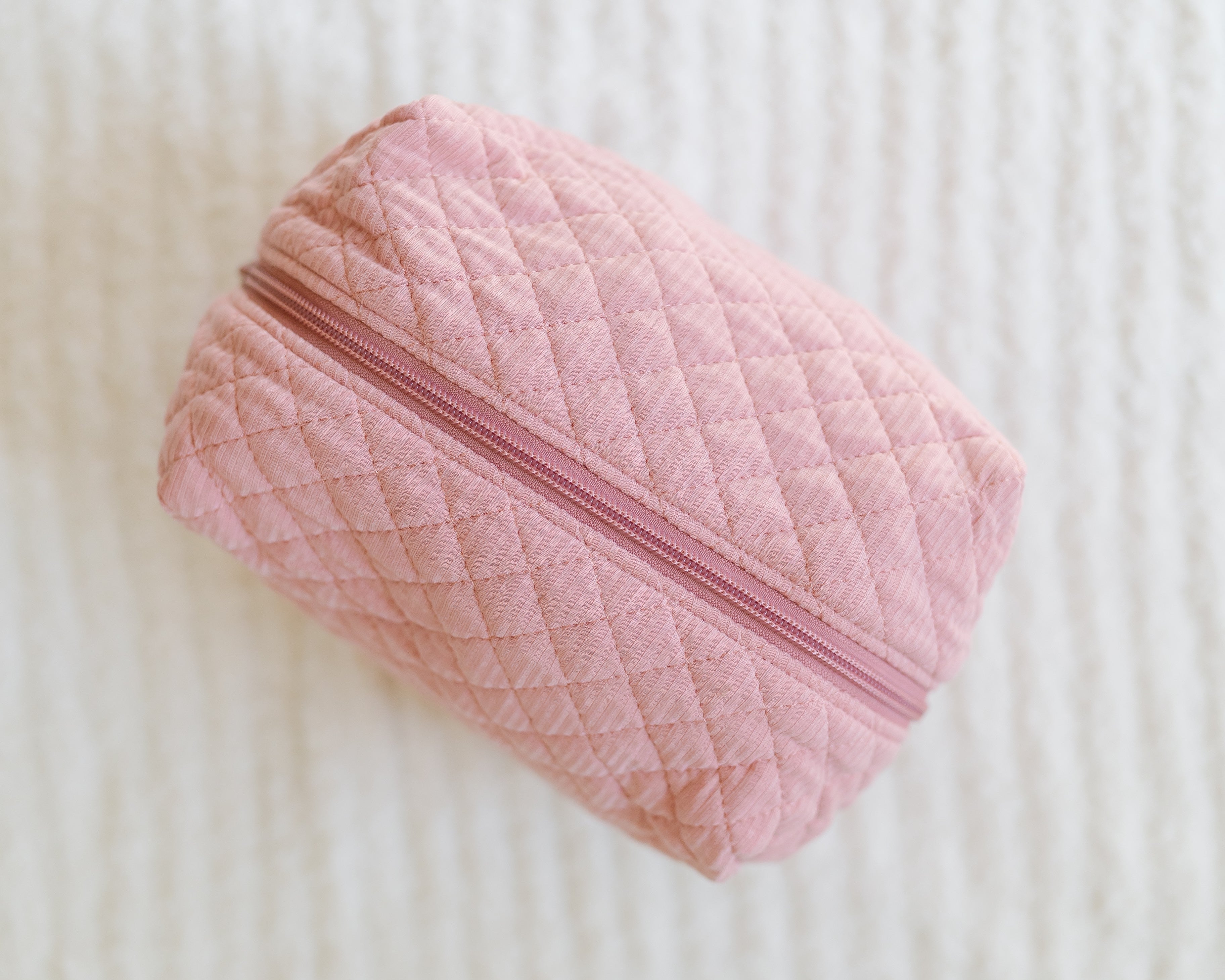A small, rectangular, quilted pink pouch with a zipper—called the peony travel bag—rests on a white textured surface, making it a cute and practical accessory for your travels.
