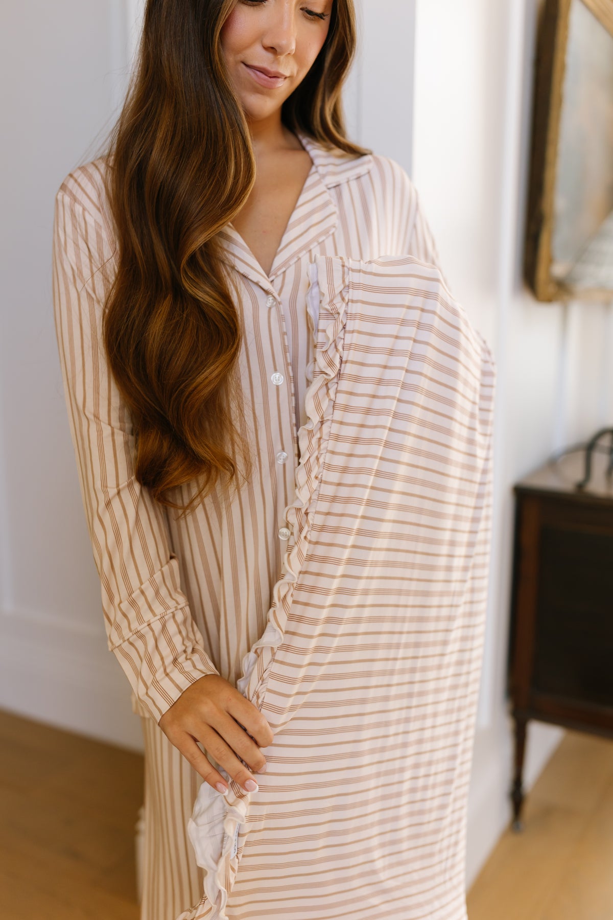 A woman with long brown hair wears the toast and honey / ruffle beige and white striped nightgown and holds a luxurious, matching striped toast and honey / ruffle blanket, standing indoors near a piece of furniture.