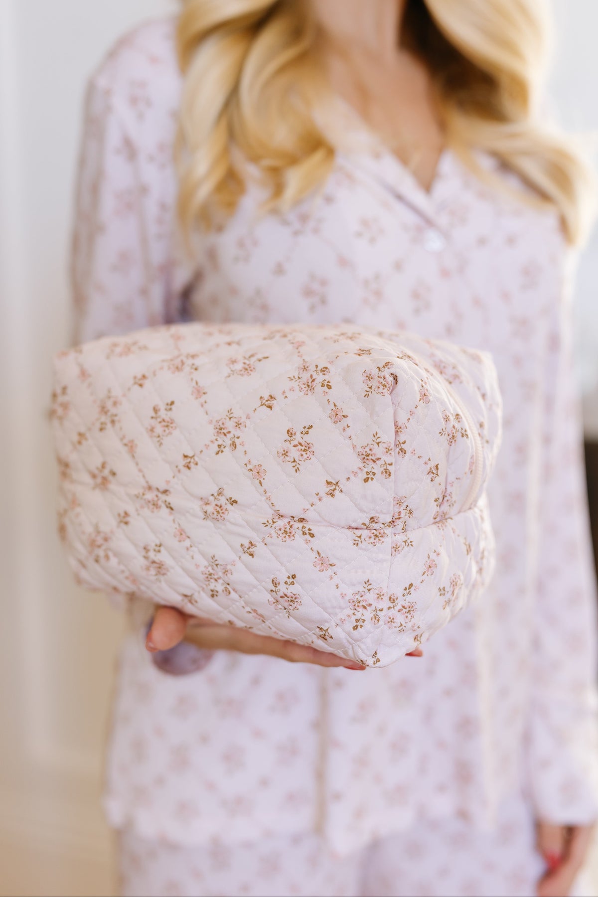 A woman in light floral pajamas holds a folded, matching keepsake / travel bag featuring subtle Goosebumps prints. Her face is not visible, and her blonde hair is styled in loose waves.