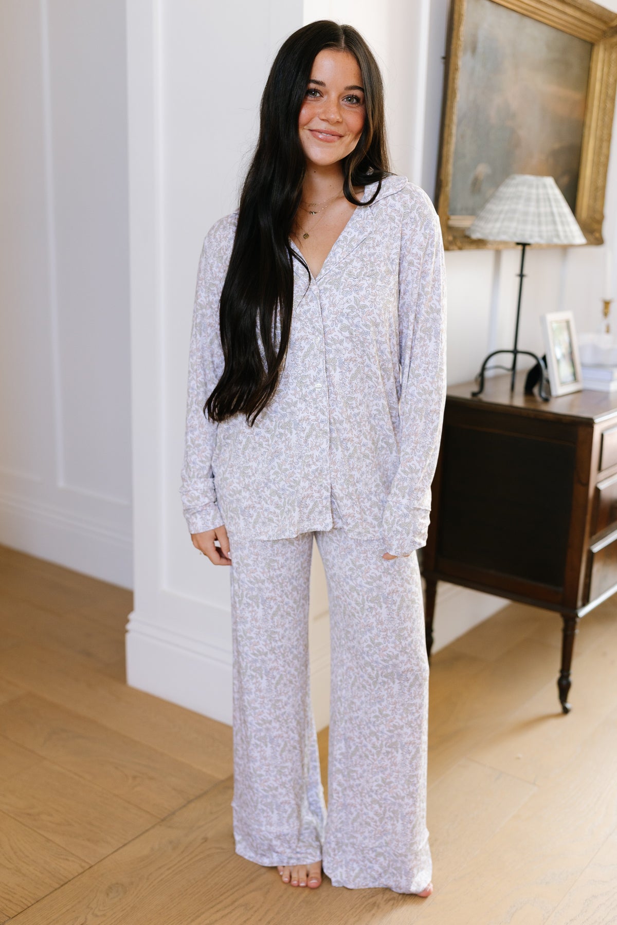 A woman with long dark hair stands indoors, smiling and wearing the heirloom pajama set with long pants in a light color. She is barefoot on a wooden floor, with a table, lamp, and framed picture in the background.