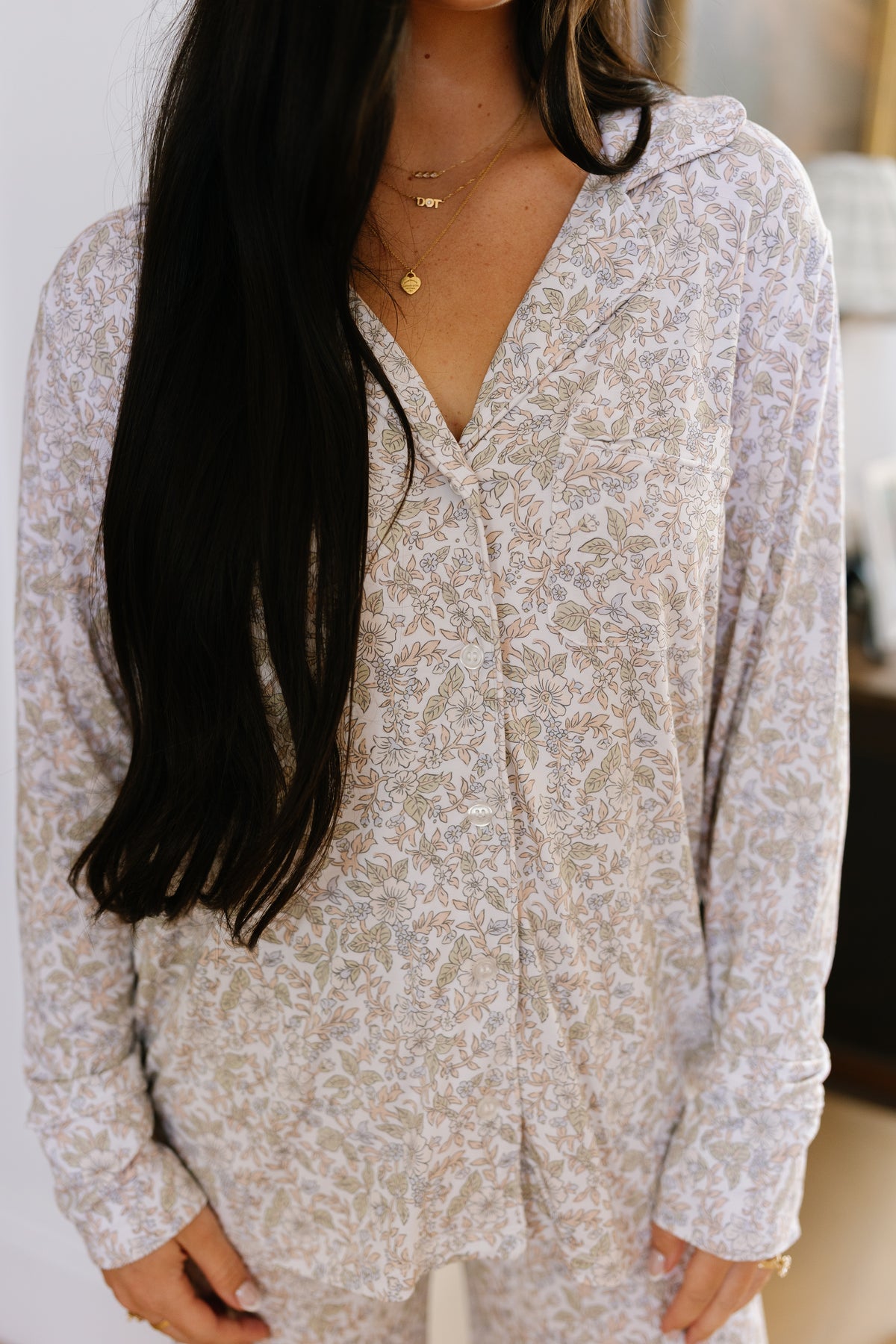 A woman with long dark hair stands indoors wearing the heirloom pajama set (long pant) in a matching pastel floral print, paired with gold necklaces. Her face is partially out of frame, drawing attention to the relaxed fit and effortless style of this loungewear sleepwear.