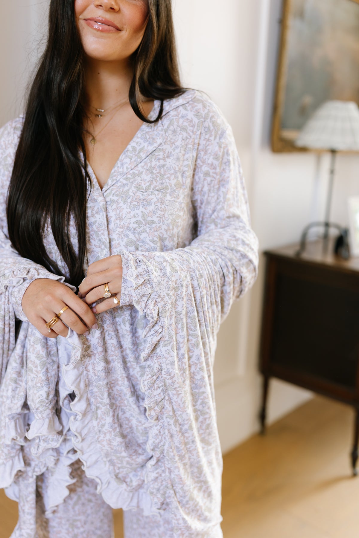 A woman with long dark hair smiles gently while wearing the heirloom pajama set with long pants, featuring a light, patterned, ruffled floral design. She accessorizes with several rings on her fingers. The background includes a wooden cabinet, lamp, and painting.