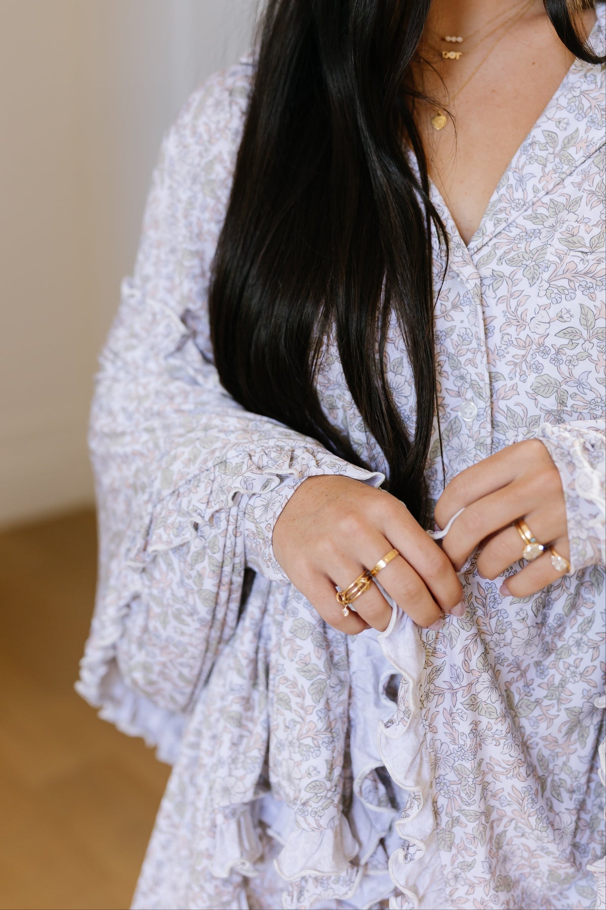 A woman with long dark hair, wearing a light floral dress and several gold rings, gently holds her hair while the heirloom / ruffle blanket softly drapes across her lap. Only her lower face and hands are visible.