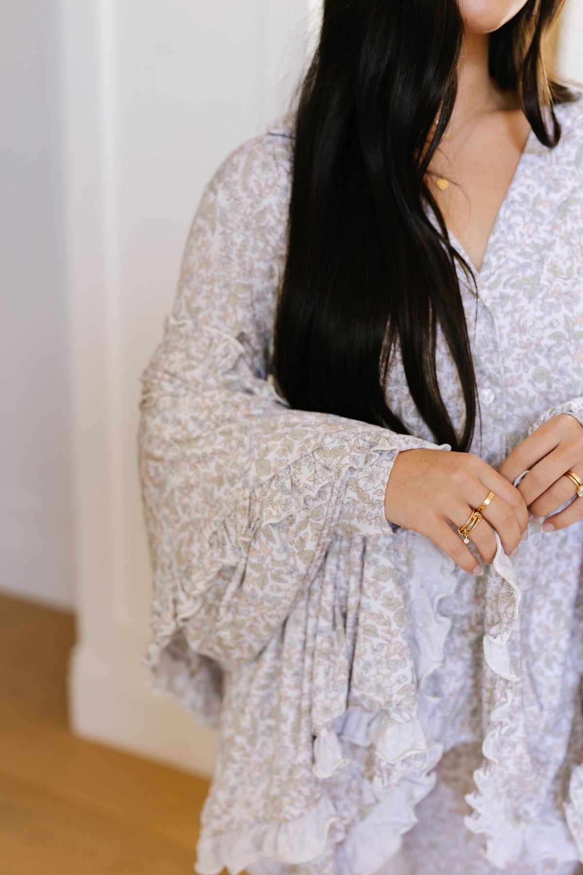 A woman with long dark hair stands indoors wearing a light floral dress with ruffled sleeves and multiple gold rings, elegantly draping the luxurious heirloom / ruffle bamboo blanket over her arm.