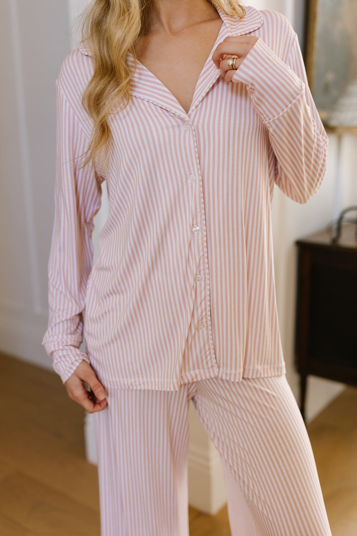 A person wearing the heritage / pajama set / long pant in pink and white stripes stands indoors with one hand in a pocket and the other touching their collar. Their face is partially out of the frame, perfectly capturing the relaxed vibe of women’s sleepwear and loungewear.