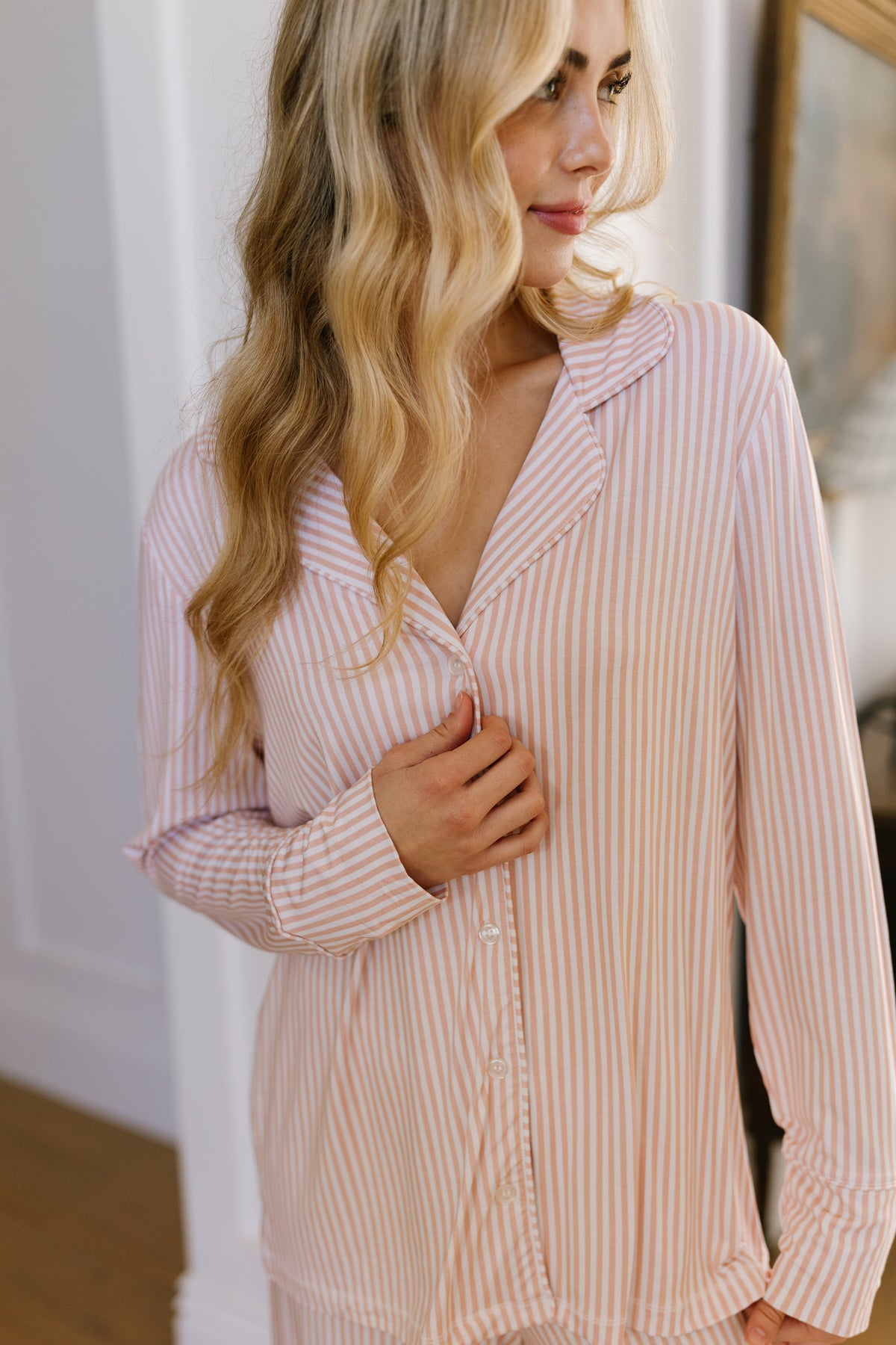 A woman with long wavy blonde hair wears the heritage pajama set with long pants, featuring pink and white stripes in soft bamboo fabric, gently holding the shirt near her chest as she stands indoors under soft light—a perfect example of comfortable women’s loungewear.