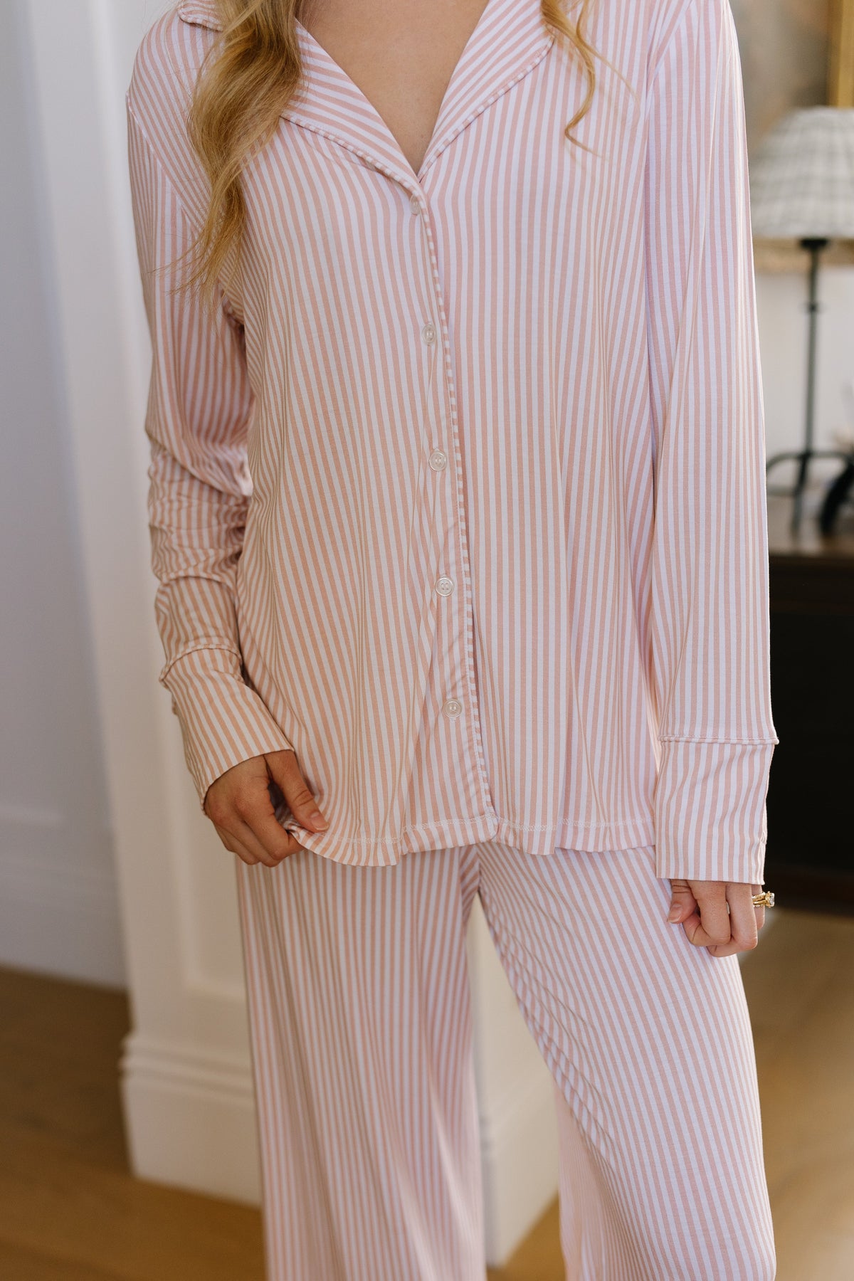 A person is seen indoors wearing the heritage pajama set / long pant in a light pink and white striped pattern, with their hand resting by their side and their face out of view.