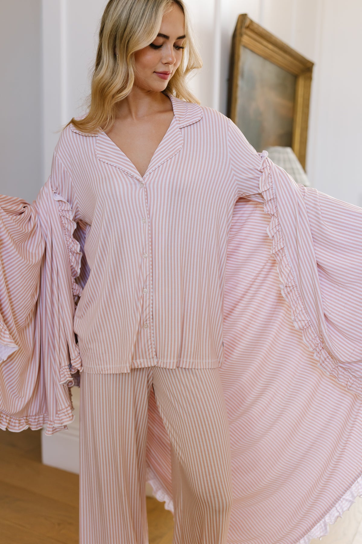 A woman stands indoors in front of a painting and white walls, holding out the sides of her heritage / ruffle pajamas to showcase their light pink and white striped fabric with ruffled, flowing sleeves. The cozy setting evokes the comfort of relaxing under a luxurious bamboo blanket.