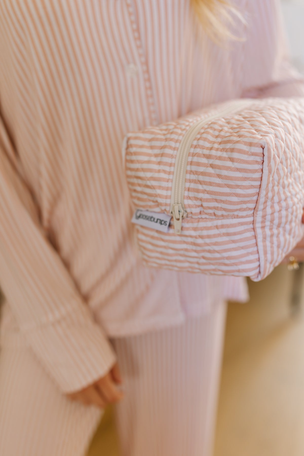Wearing pink and white striped pajamas, a person holds the heritage / travel bag—a matching zippered pouch ideal for organizing travel accessories—with a small tag that reads stoneyclover.