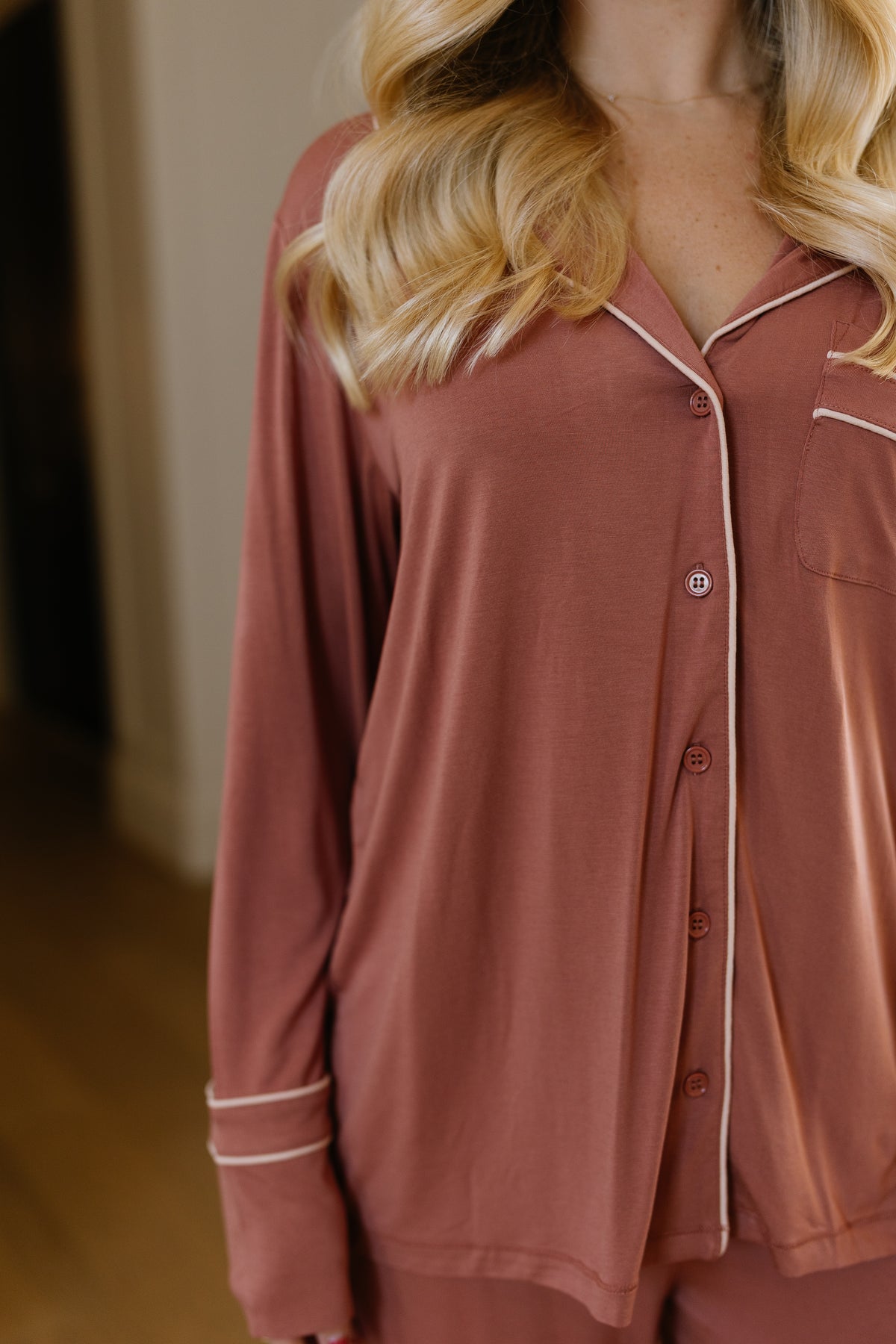 A woman with long blonde hair is seen indoors wearing the serenity pajama set with long pants in a relaxed-fit, mauve satin design. Her face is not shown, as the image highlights the upper body and details of the pajama top.