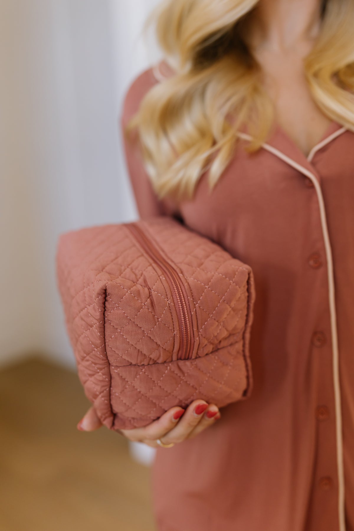 A woman in a pink robe holds a quilted, square pink serenity / travel bag zipper pouch in one hand. The focus is on the pouch—an essential among stylish travel accessories—with her face out of frame and her blonde hair visible.