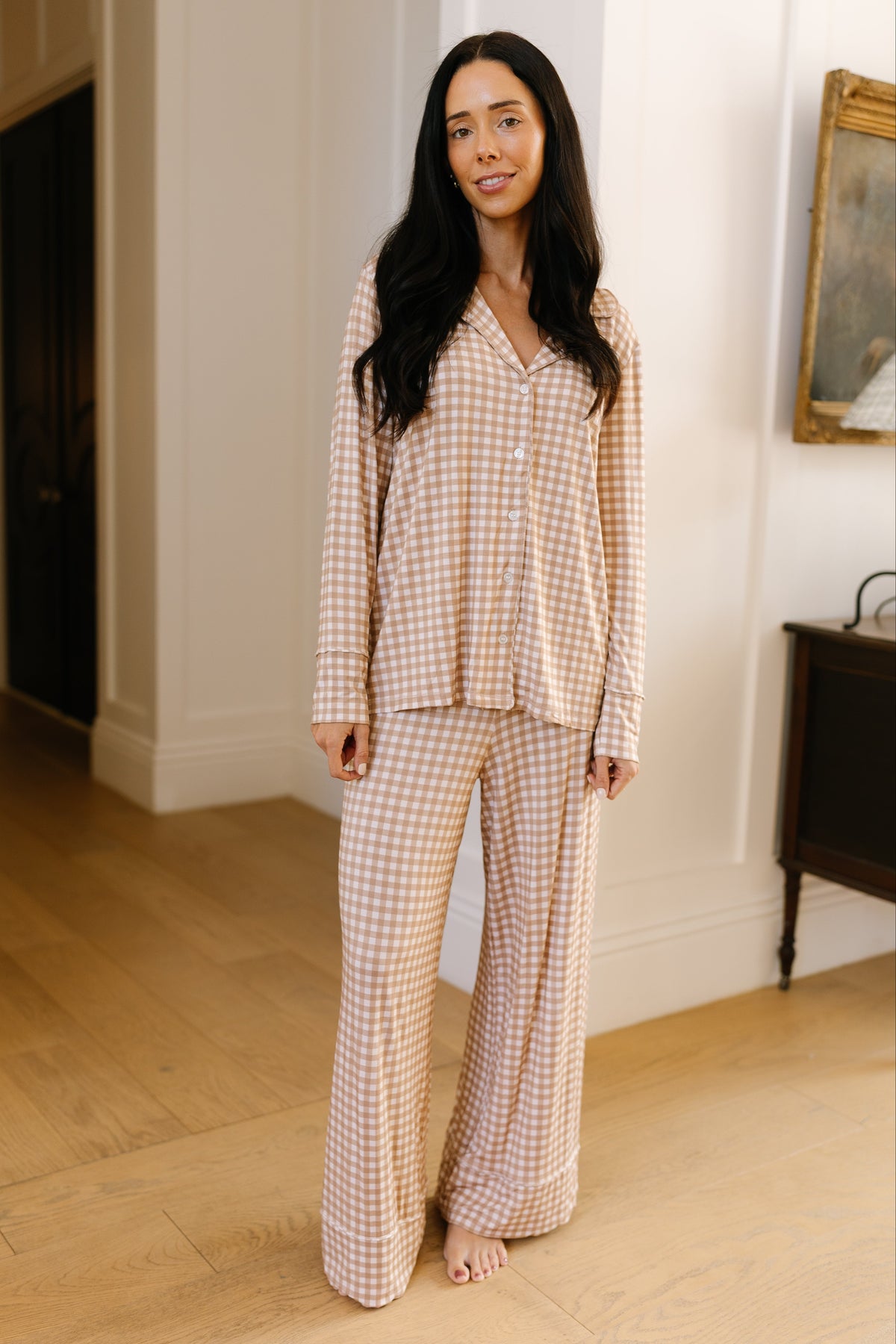 A woman with long dark hair stands indoors, barefoot, smiling at the camera while wearing the teddy bear check pajama set with long pants in a relaxed beige and white checked pattern.