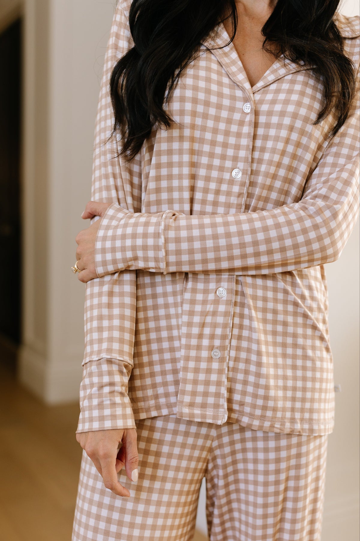 A woman stands indoors with her arms gently crossed in front of her, wearing the teddy bear check pajama set with long pants—a relaxed-fit loungewear ensemble in a beige and white gingham pattern. The timeless design features long sleeves, and her face is partly out of the frame.
