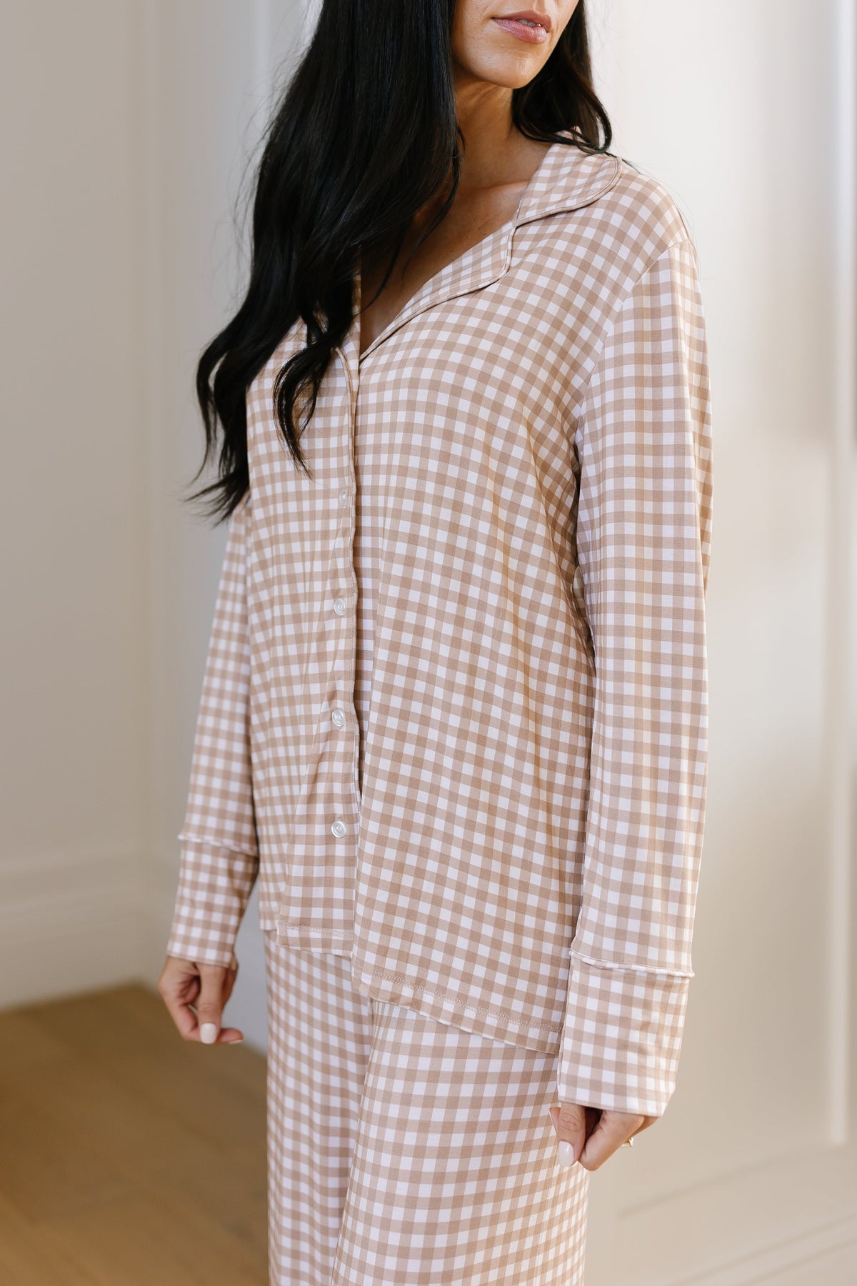 A woman wearing the teddy bear check pajama set with long pants stands indoors, facing slightly sideways. Her long dark hair is down, and only her torso and part of her face are visible. The relaxed fit of the pajamas is highlighted in a softly lit room, enhancing the cozy atmosphere.