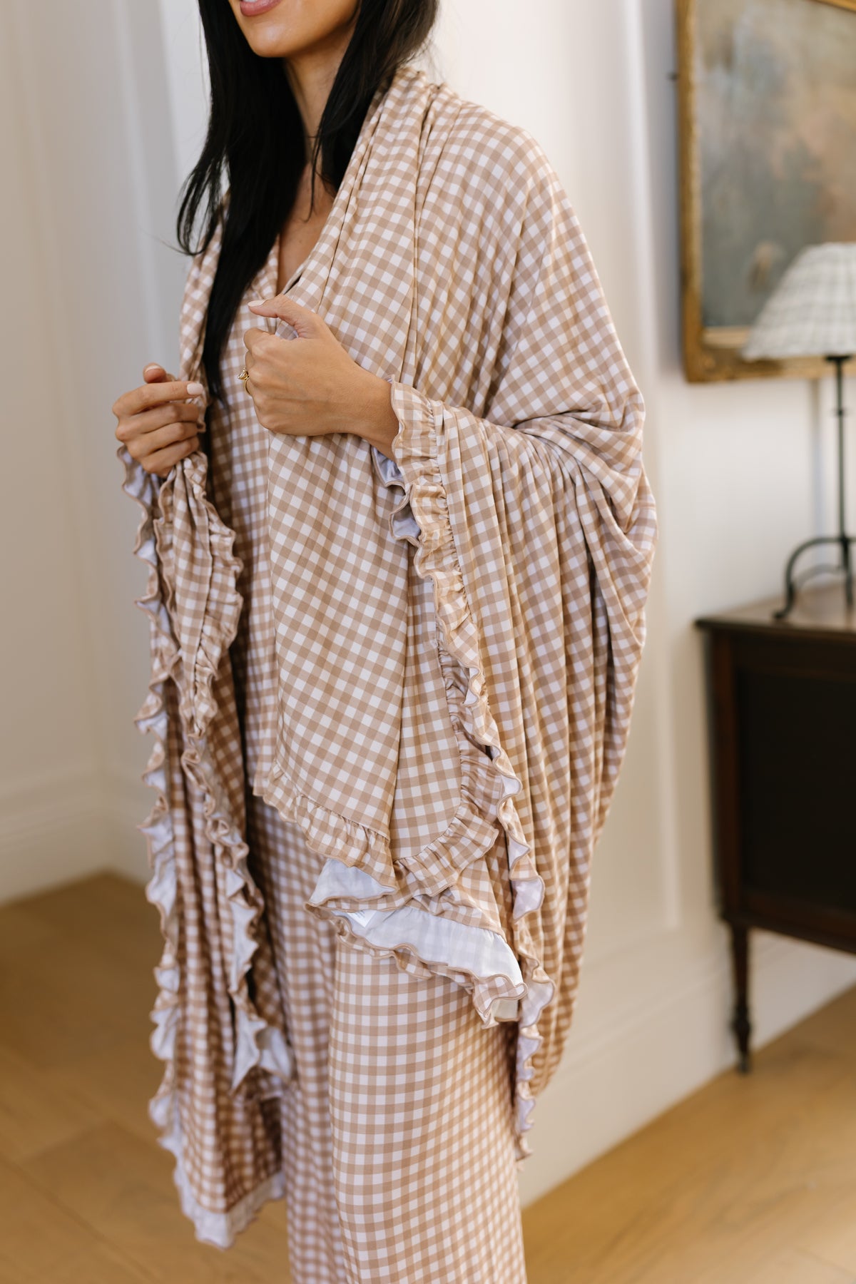 A woman stands indoors, partially out of frame, wearing the teddy bear check / ruffle dress and holding its matching luxurious blanket-like ruffled shawl around her shoulders.