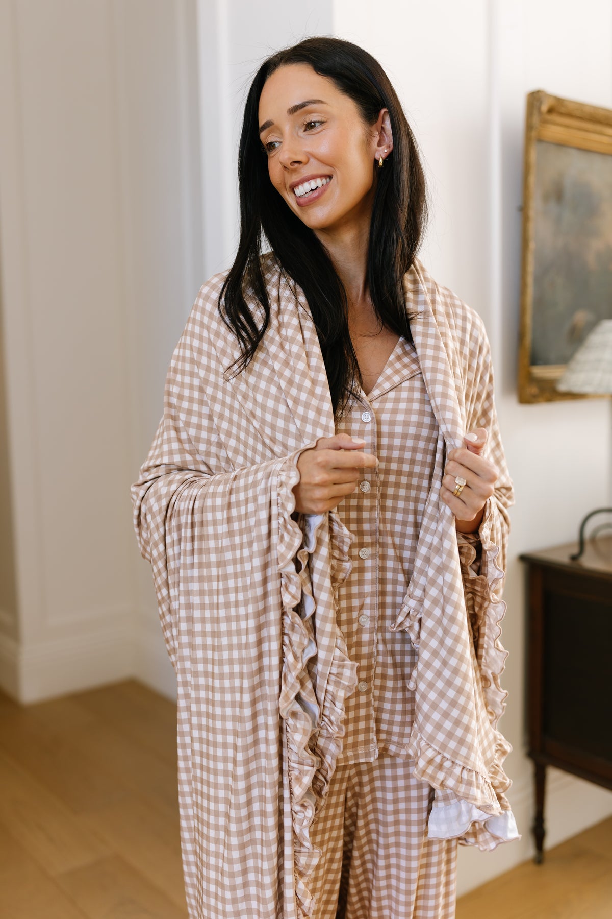 A woman smiles while standing indoors near a painting and lamp, wearing the “teddy bear check / ruffle” pajamas and a matching ruffled shawl, her look complemented by a soft “teddy bear check / ruffle” blanket draped nearby.