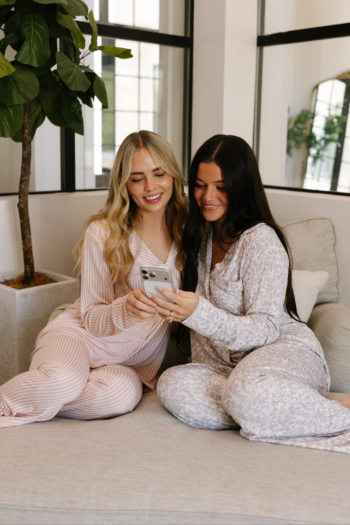 Two women wearing the heirloom pajama set with long pants sit on a couch, smiling and looking at a smartphone together. A large potted plant and a window are visible in the background.