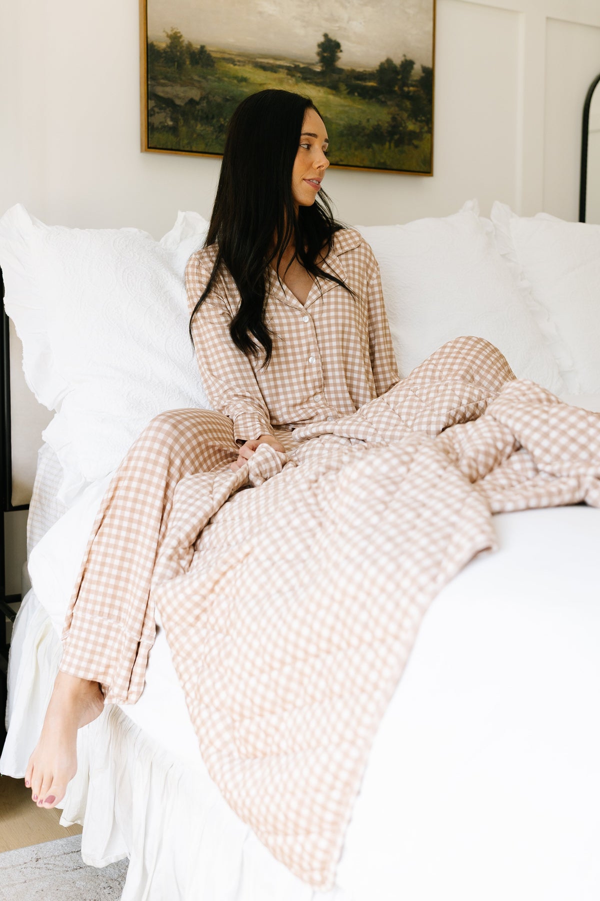 A woman in the teddy bear check pajama set with long pants sits on a bed with white bedding, holding a matching blanket. She looks to the side, with a painting and white pillows behind her, perfectly showcasing the relaxed fit of this cozy loungewear—ideal for comfortable evenings at home.