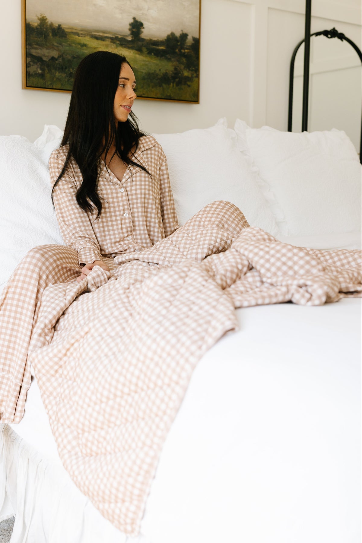 A woman wearing beige teddy bear check pajamas sits on a white bed, gazing to the side. She is wrapped in a teddy bear check quilt that matches her pajamas. The bright room features white bedding and a landscape painting on the wall behind her.