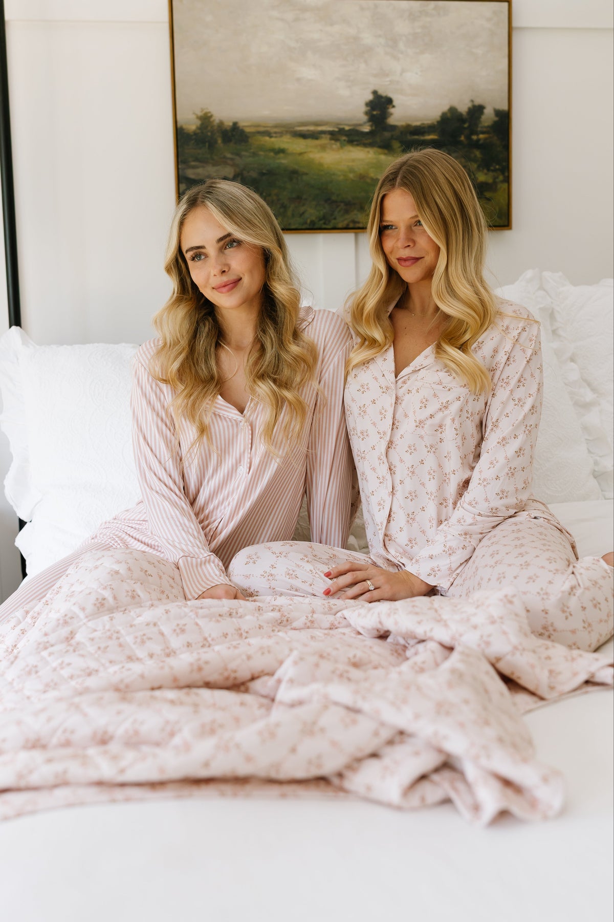 Two women with long blonde hair sit close together on a bed, smiling as they wrap themselves in the keepsake / quilt. They wear light pink patterned pajamas, and a landscape painting hangs on the wall behind them.