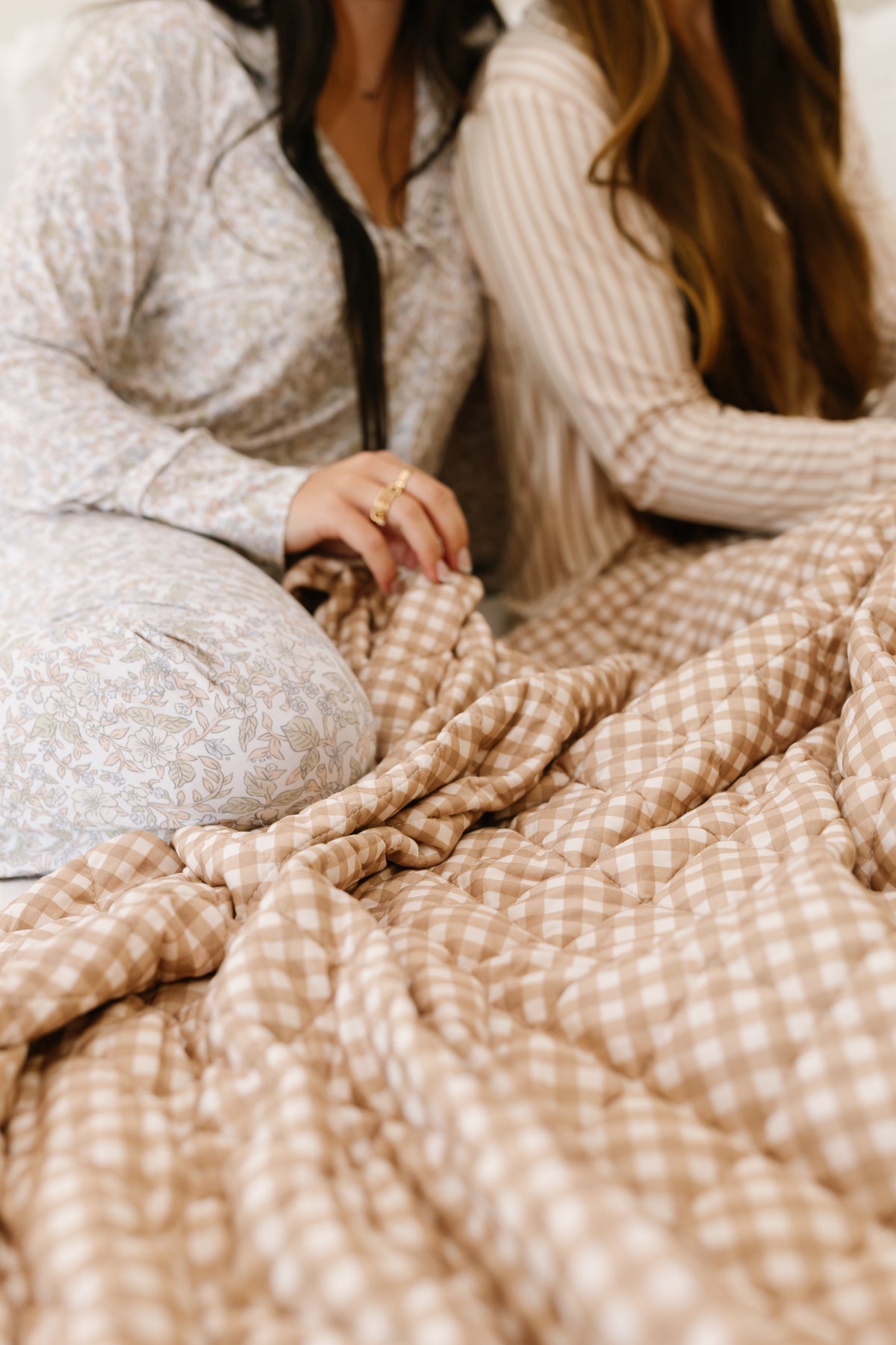 Two women in patterned pajamas sit closely together under a beige and white teddy bear check quilt. Their faces are not visible, drawing attention to their clothing and the cozy teddy bear check bedding.