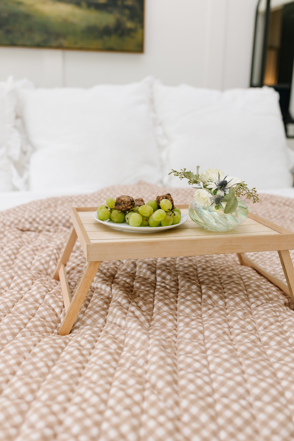 A teddy bear check quilt is softly folded on the bed, topped with a wooden breakfast tray displaying a plate of green grapes and pastries beside a small vase of white flowers.