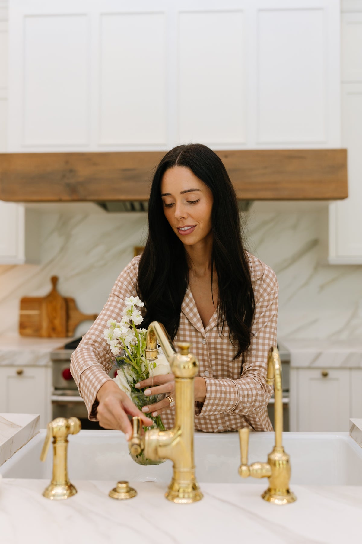 A woman with long dark hair, wearing the teddy bear check pajama set with long pants, fills a glass vase of white flowers with water at a kitchen sink featuring gold fixtures and a marble backsplash.