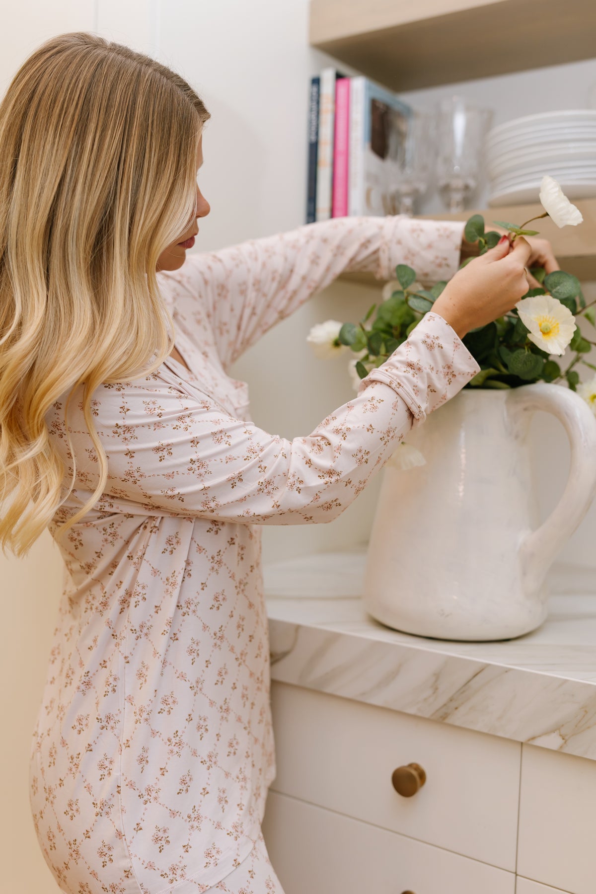 A woman with long blonde hair, dressed in the keepsake pajama set with long pants, arranges white flowers in a large white pitcher on a marble kitchen counter, with shelves holding books and glassware in the background.
