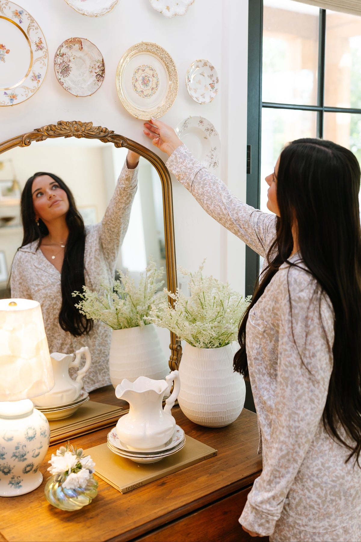Wearing the heirloom long pant pajama set, a woman in a floral ensemble reaches up to adjust a decorative plate on the wall above a wooden dresser adorned with a mirror, vase, lamp, and ceramic pitcher—an inviting scene that perfectly complements the cozy feel of women’s loungewear.