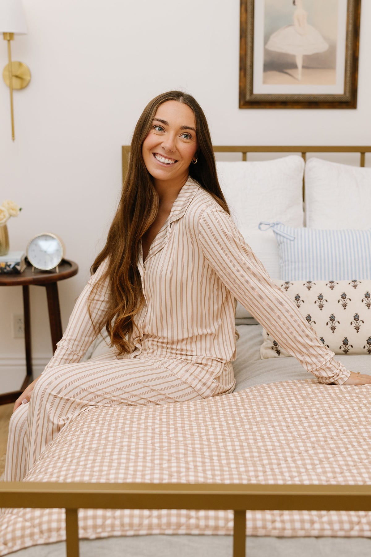 A woman with long brown hair, wearing the toast and honey pajama set in a relaxed fit with beige and white stripes, sits smiling on a bed adorned with patterned pillows in a cozy, well-decorated bedroom.