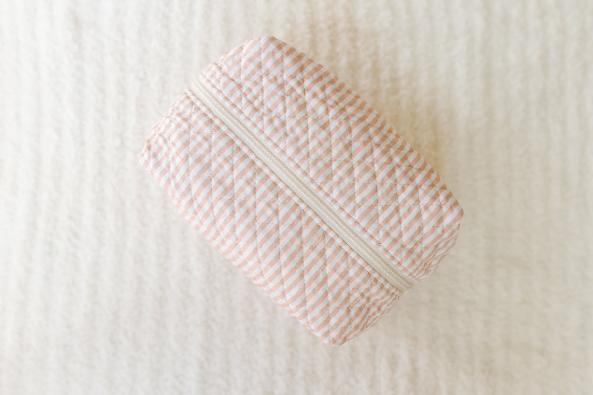 Rectangular fabric heritage / travel bag featuring pink and white diagonal stripes with a white zipper, displayed on a soft, textured cream-colored surface. Perfect for keeping your travel accessories organized in style.