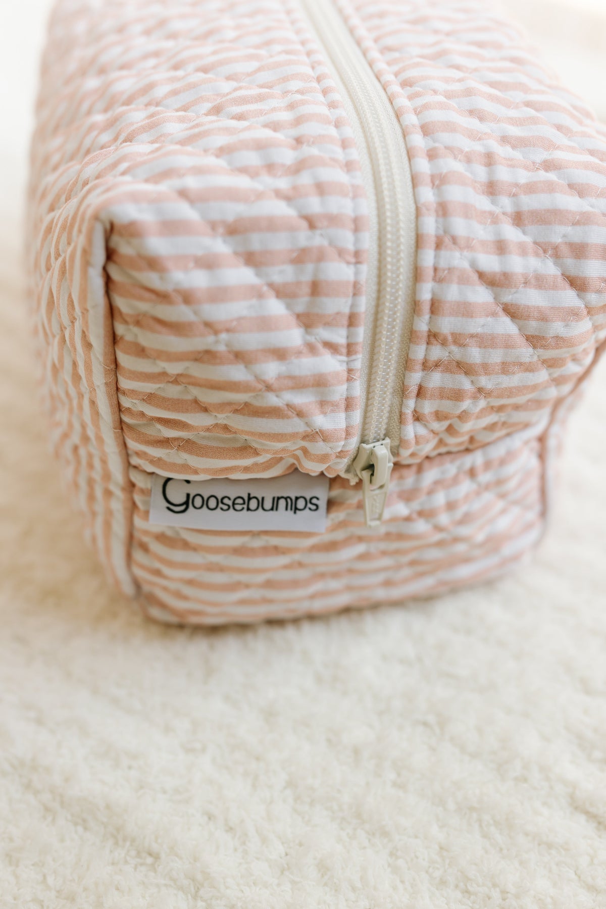 A quilted, rectangular pouch with pink and white stripes, a white zipper, and a Goosebumps label rests on a soft, cream-colored textured surface—this heritage / travel bag is an essential accessory for organizing your travel essentials.