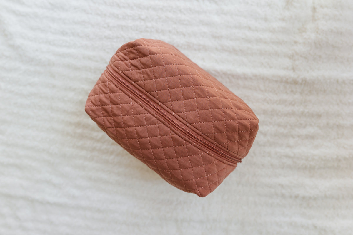 A quilted, rectangular serenity / travel bag in dusty rose with a zipper sits atop a soft, white textured surface—an elegant addition to your travel accessories collection.