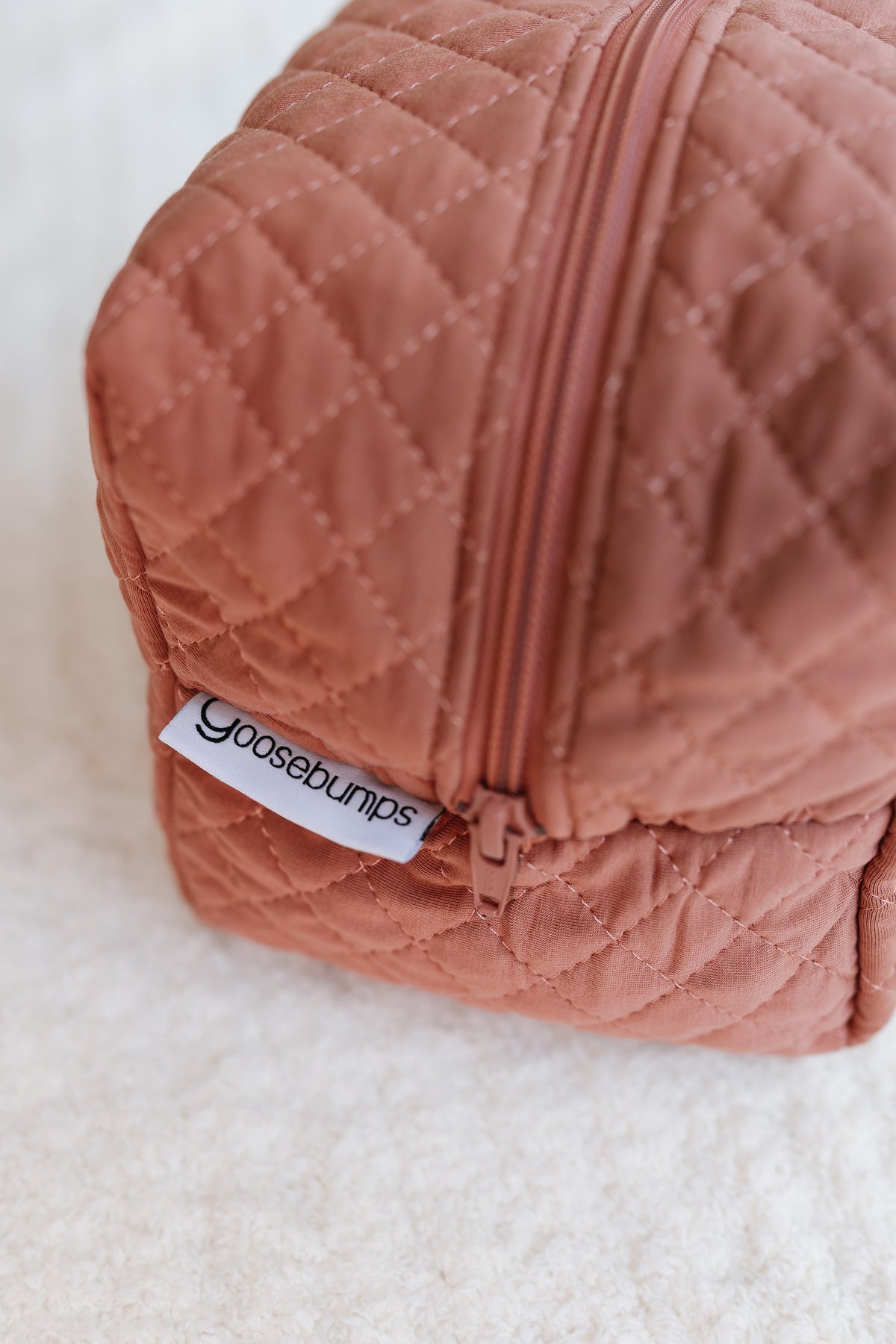 A close-up of a quilted, rust-colored serenity / travel bag with a zipper and a white tag labeled goosebumps, placed on a soft, white textured surface—an ideal choice for those seeking stylish travel accessories.