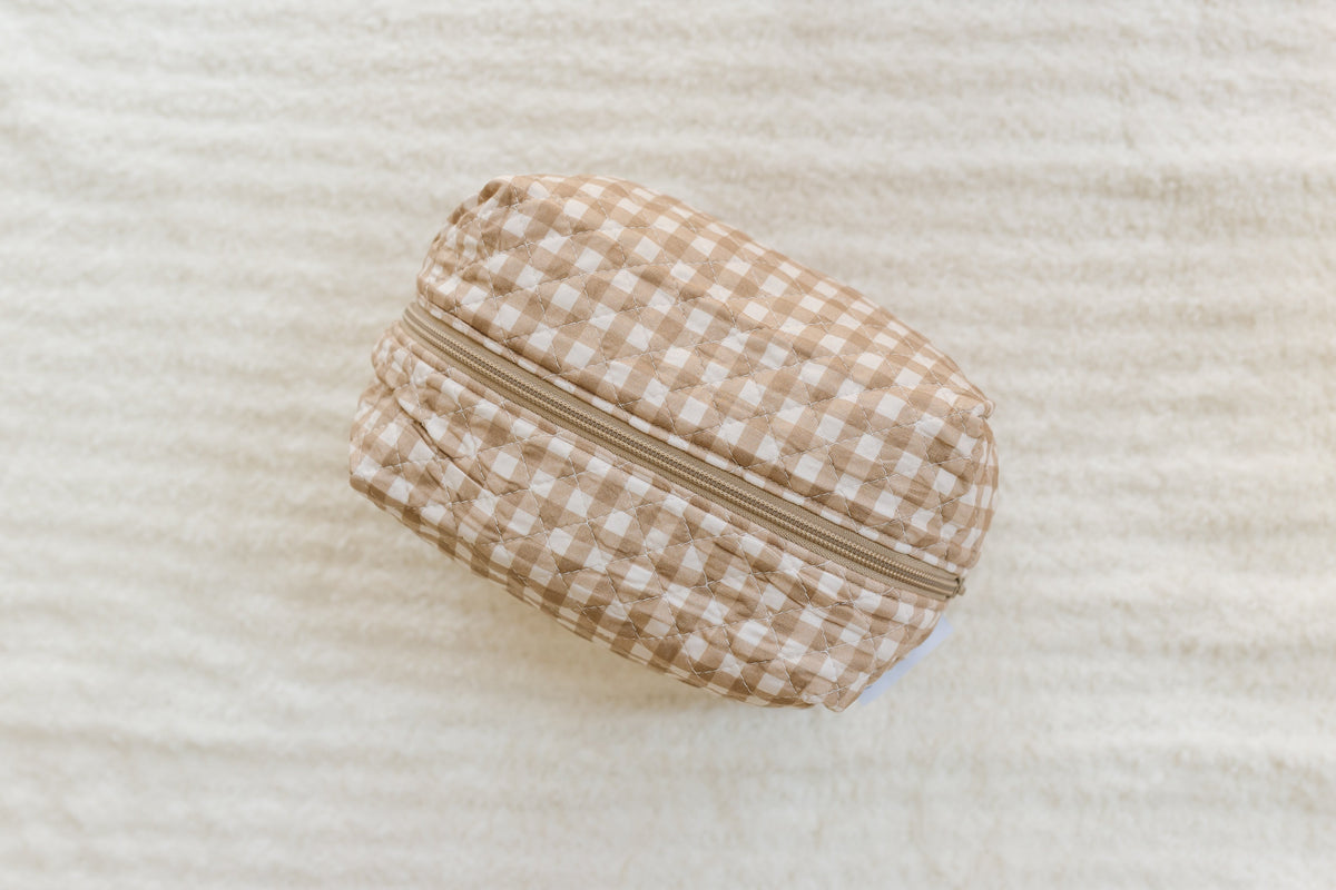 A teddy bear check travel bag—a beige and white gingham zippered pouch—rests on a soft, textured cream-colored surface, viewed from above.
