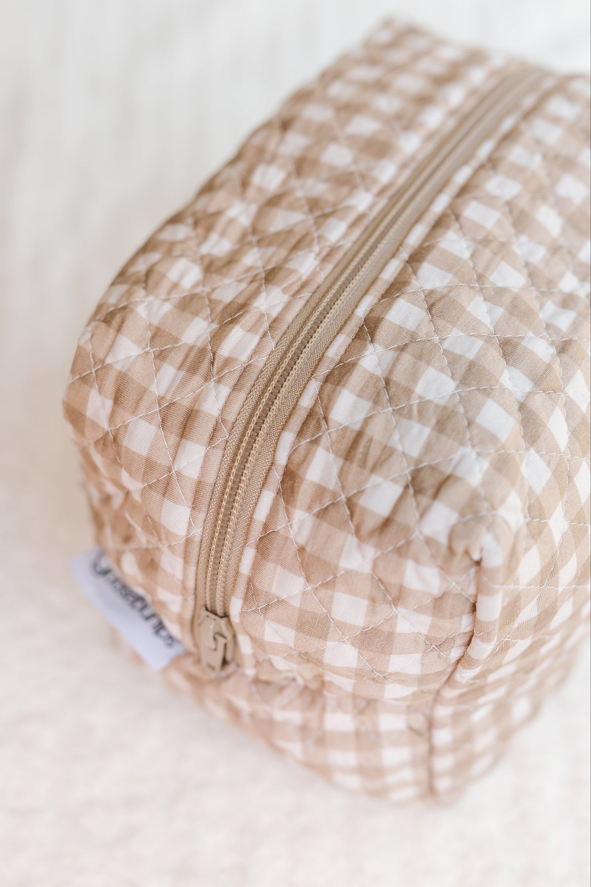 A quilted, beige and white checkered zippered pouch—named the teddy bear check / travel bag—rests on a soft, cream-colored surface. A small fabric tag accents the side of the pouch, enhancing its charming design as an elegant addition to your travel accessories collection. Perfect as a Goosebumps Travel Bag, it seamlessly blends style and function for journeys both near and far.