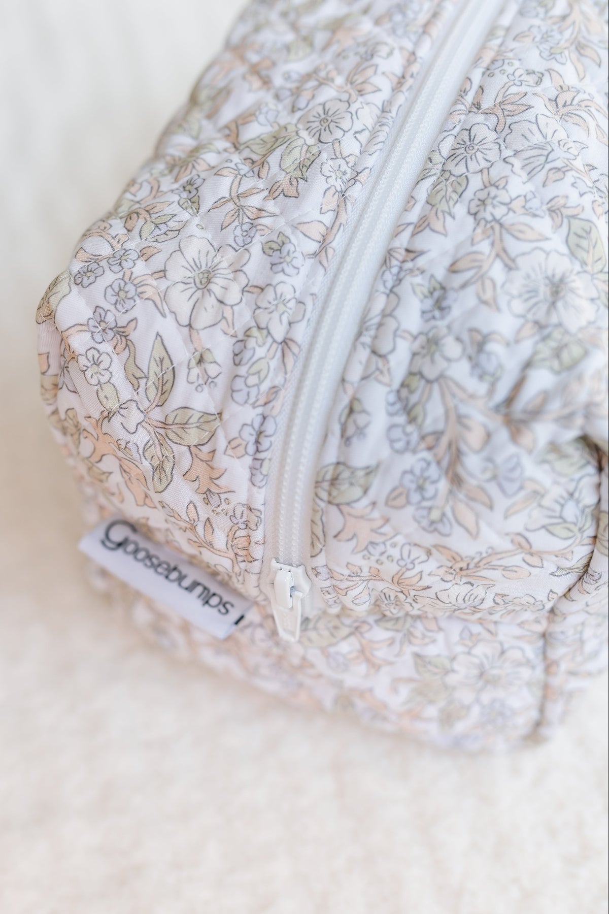 A close-up of a quilted fabric pouch adorned with a floral pattern and finished with a white zipper. A small label reading “Gooselings” is attached near the zipper, highlighting its stylish design as an heirloom / travel bag. The pouch rests on a soft, cream-colored surface.