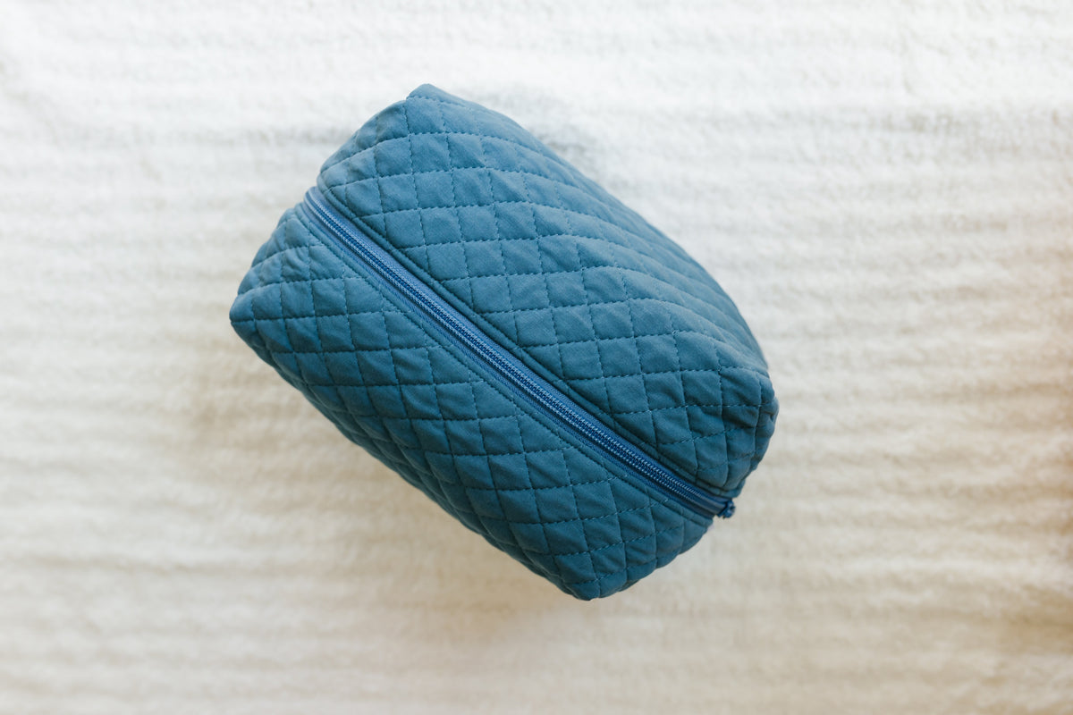A stylish blue quilted “tidal wave / travel bag” with a zipper rests on a textured white surface, making it the perfect addition to your travel accessories collection.