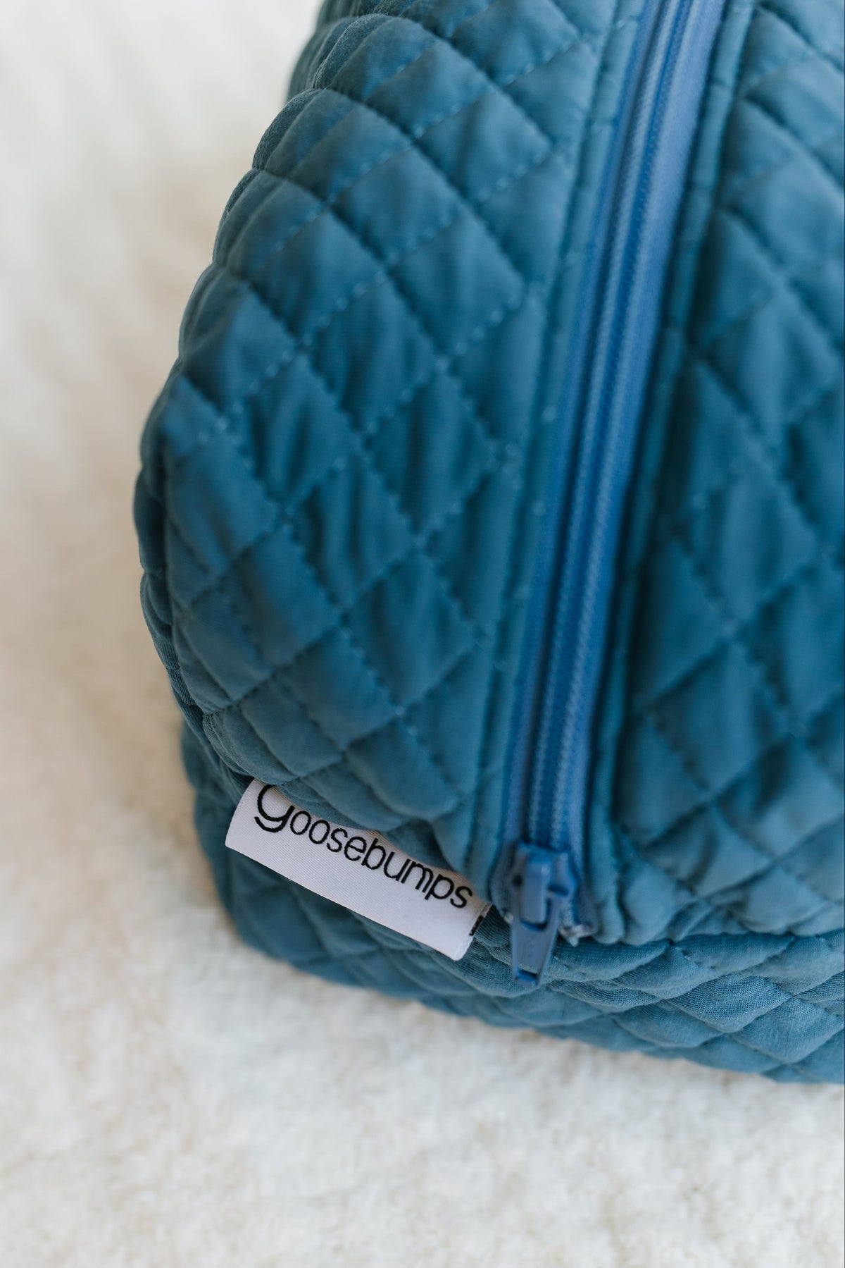 A close-up of a blue quilted tidal wave / travel bag with a visible zipper and a white tag labeled Goosebumps, lying on a soft, white textured surface—an essential addition to your travel accessories collection.