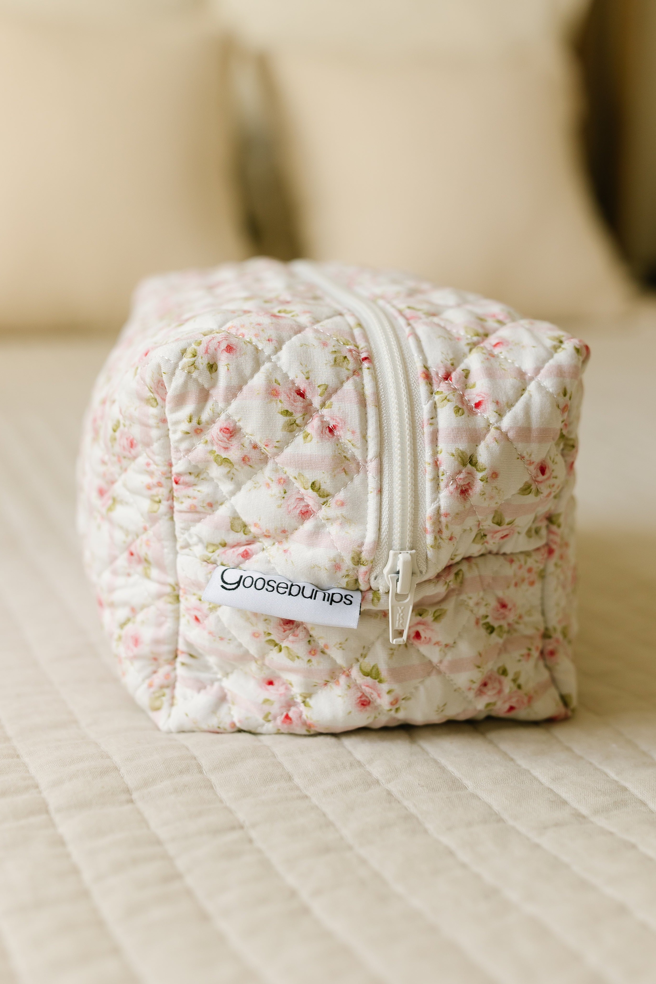 The rosie / travel bag, a quilted floral-patterned Goosebumps bag with zipper and tag, sits on a tidy bed with cream pillows and a wooden headboard—an elegant upgrade for your travel accessories collection.